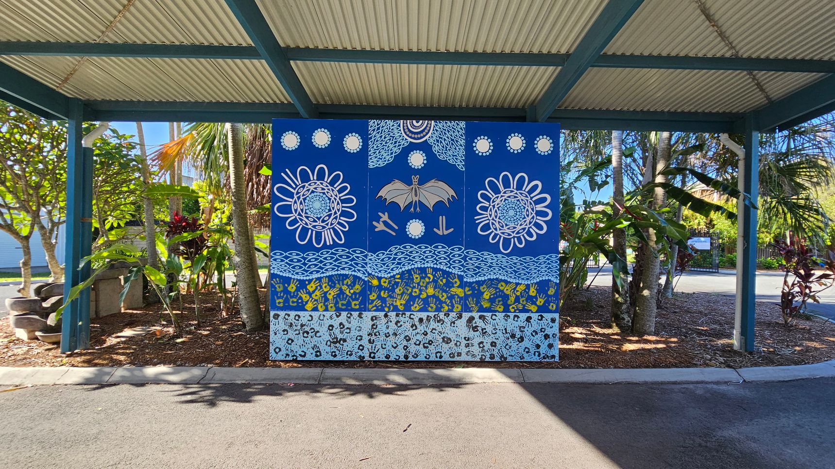 A vibrant mural is centred under a corrugated metal roof supported by green beams. The mural features a blue background with intricate designs, including a bat, circular patterns, and handprints in yellow and black. Surrounding the mural are lush tropical plants with broad leaves and some red foliage. The ground is covered with mulch and the foreground has a paved area. A vibrant mural is centred under a corrugated metal roof supported by green beams. The mural features a blue background with intricate designs, including a bat, circular patterns, and handprints in yellow and black. Surrounding the mural are lush tropical plants with broad leaves and some red foliage. The ground is covered with mulch and the foreground has a paved area.
