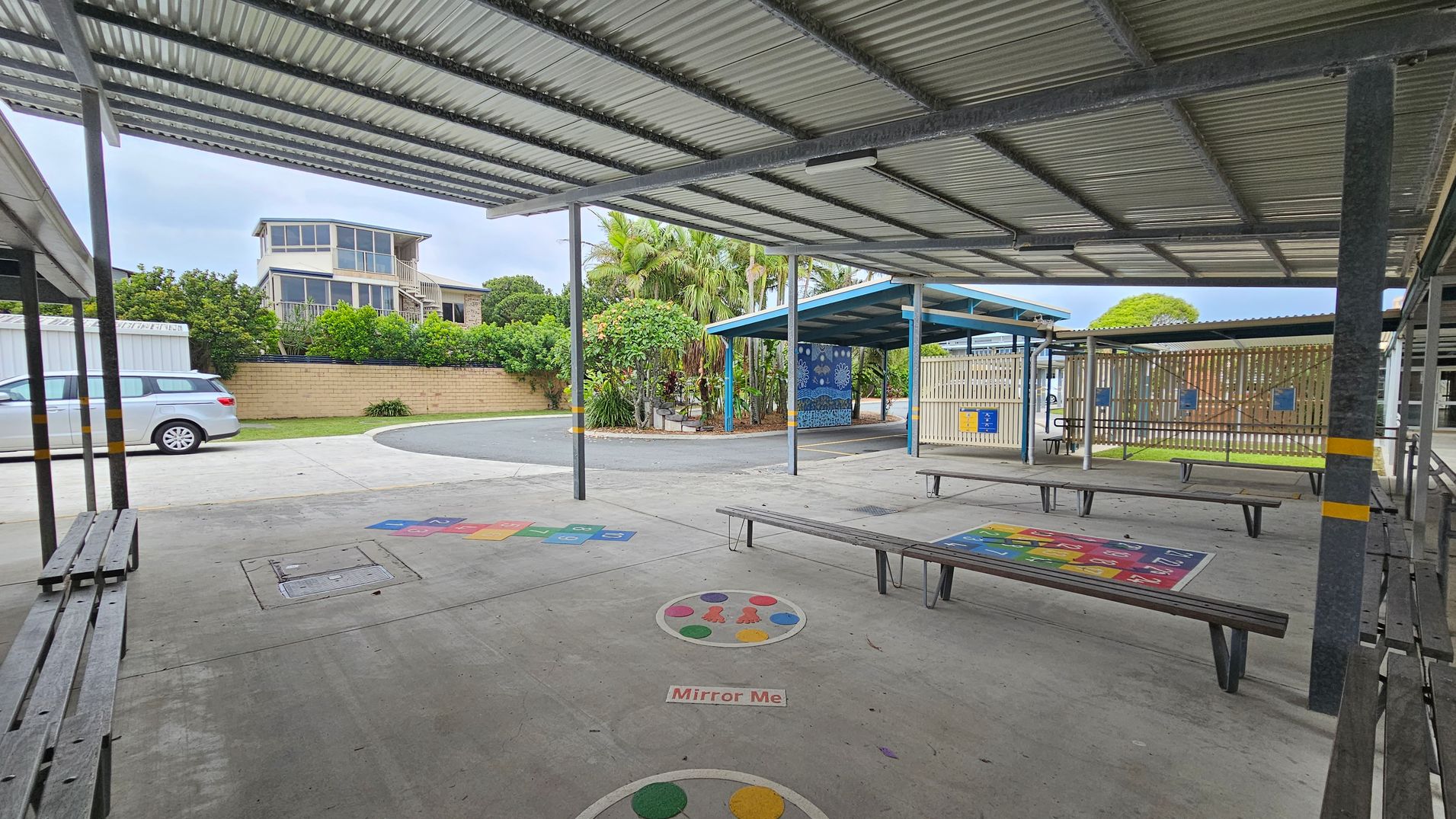 A covered outdoor area with metal roofing and benches. The concrete floor has colourful game markings, including a "Mirror Me" circle. In the background, there's a two-storey building with large windows, a parked silver car, and a brick wall with lush greenery. To the right, a fenced area with a blue-roofed shelter and more benches. Palm trees and other plants are visible near the centre. A covered outdoor area with metal roofing and benches. The concrete floor has colourful game markings, including a "Mirror Me" circle. In the background, there's a two-storey building with large windows, a parked silver car, and a brick wall with lush greenery. To the right, a fenced area with a blue-roofed shelter and more benches. Palm trees and other plants are visible near the centre.