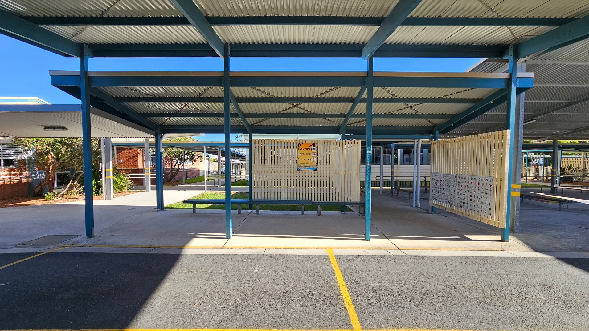 A covered outdoor area with a corrugated metal roof supported by blue metal beams. In the centre, a wooden slatted partition displays a poster. The ground is a mix of concrete and a textured surface with yellow lines. To the left, there are plants and a brick building with windows. The right side shows more covered walkways and benches. The background includes additional structures and greenery. A covered outdoor area with a corrugated metal roof supported by blue metal beams. In the centre, a wooden slatted partition displays a poster. The ground is a mix of concrete and a textured surface with yellow lines. To the left, there are plants and a brick building with windows. The right side shows more covered walkways and benches. The background includes additional structures and greenery.