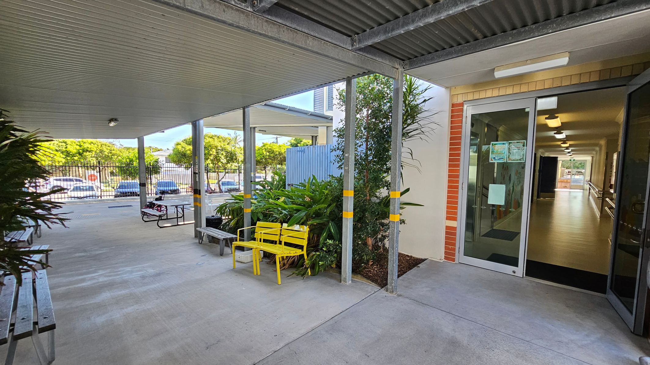A covered outdoor area with a concrete floor and a corrugated metal ceiling. On the right, there is a glass door leading to a hallway with wooden flooring and ceiling lights. Near the door, there are green plants and a yellow bench. On the left, there are metal benches and a picnic table. In the background, a fence and parked cars are visible, along with trees. A covered outdoor area with a concrete floor and a corrugated metal ceiling. On the right, there is a glass door leading to a hallway with wooden flooring and ceiling lights. Near the door, there are green plants and a yellow bench. On the left, there are metal benches and a picnic table. In the background, a fence and parked cars are visible, along with trees.