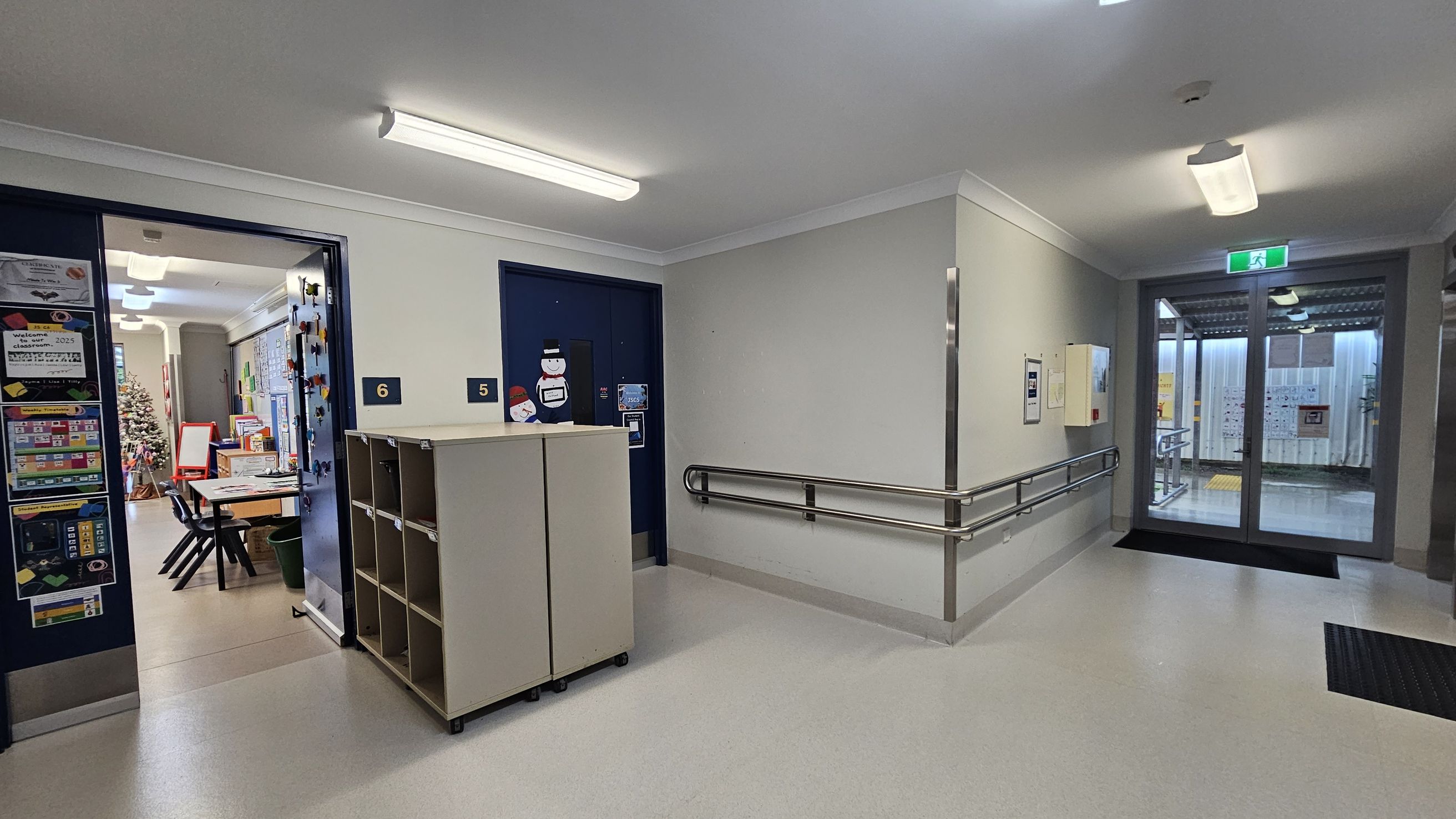 A hallway with a light grey floor and white ceiling featuring fluorescent lights. On the left, an open door leads to a classroom with tables, chairs, and a Christmas tree. The door is marked with the number 6. Adjacent is a closed door with the number 5, decorated with snowman cut-outs. A small shelving unit is positioned between the doors. On the right, a handrail runs along the wall leading to a glass door with an exit sign above, opening to an outdoor area. A hallway with a light grey floor and white ceiling featuring fluorescent lights. On the left, an open door leads to a classroom with tables, chairs, and a Christmas tree. The door is marked with the number 6. Adjacent is a closed door with the number 5, decorated with snowman cut-outs. A small shelving unit is positioned between the doors. On the right, a handrail runs along the wall leading to a glass door with an exit sign above, opening to an outdoor area.
