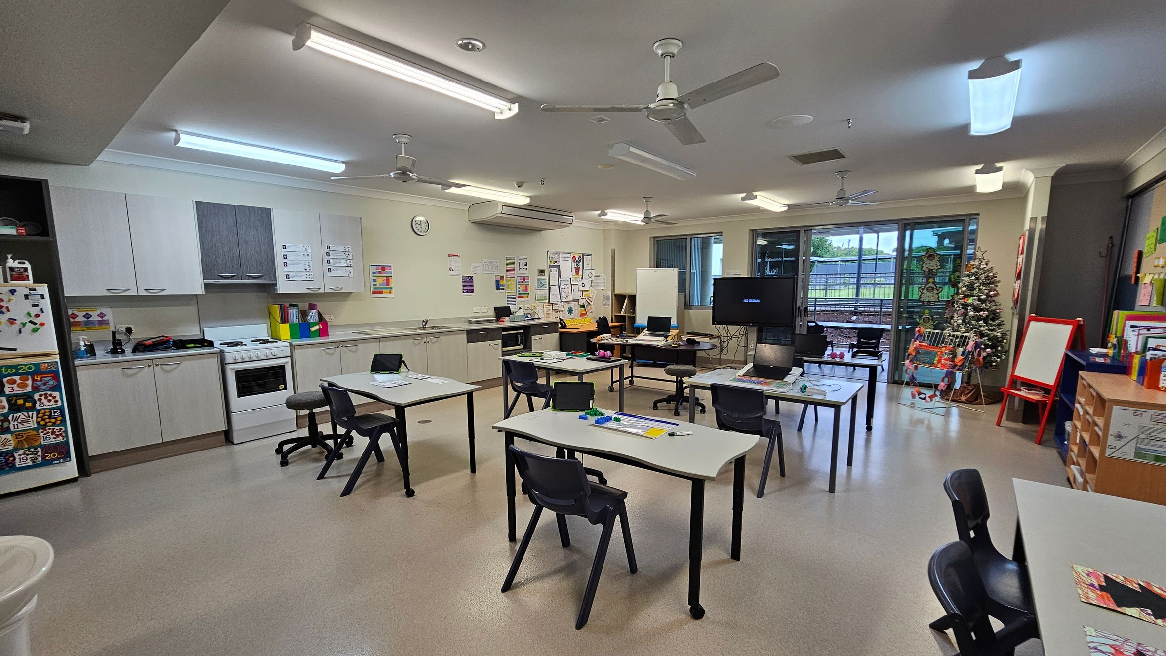 A classroom with a kitchen area on the left, featuring white cabinets, a stove, and a fridge adorned with colourful magnets. In the centre, several tables and chairs are arranged, some with laptops and educational materials. The right side has a Christmas tree and a red easel. The back wall has large windows and a door leading outside. The ceiling has fluorescent lights and ceiling fans, and the floor is a light-coloured linoleum. A classroom with a kitchen area on the left, featuring white cabinets, a stove, and a fridge adorned with colourful magnets. In the centre, several tables and chairs are arranged, some with laptops and educational materials. The right side has a Christmas tree and a red easel. The back wall has large windows and a door leading outside. The ceiling has fluorescent lights and ceiling fans, and the floor is a light-coloured linoleum.