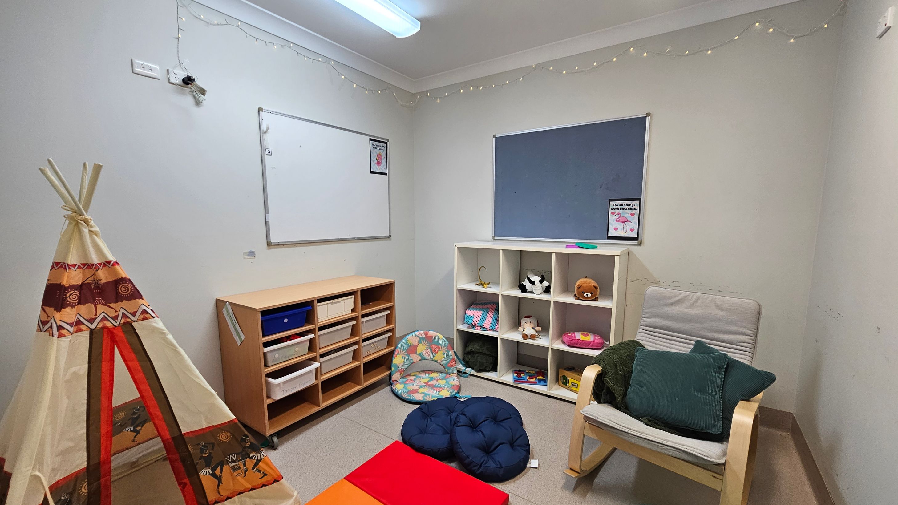 A small room with a teepee on the left, decorated with patterns in brown, orange, and red. In the centre, a wooden shelf unit with various storage boxes. To the right, a white cubby shelf holds toys and books. A cushioned chair with green pillows is nearby. The walls have a whiteboard and a blue notice board. The ceiling has a fluorescent light and string lights along the edges. The floor is carpeted in a neutral tone. A small room with a teepee on the left, decorated with patterns in brown, orange, and red. In the centre, a wooden shelf unit with various storage boxes. To the right, a white cubby shelf holds toys and books. A cushioned chair with green pillows is nearby. The walls have a whiteboard and a blue notice board. The ceiling has a fluorescent light and string lights along the edges. The floor is carpeted in a neutral tone.