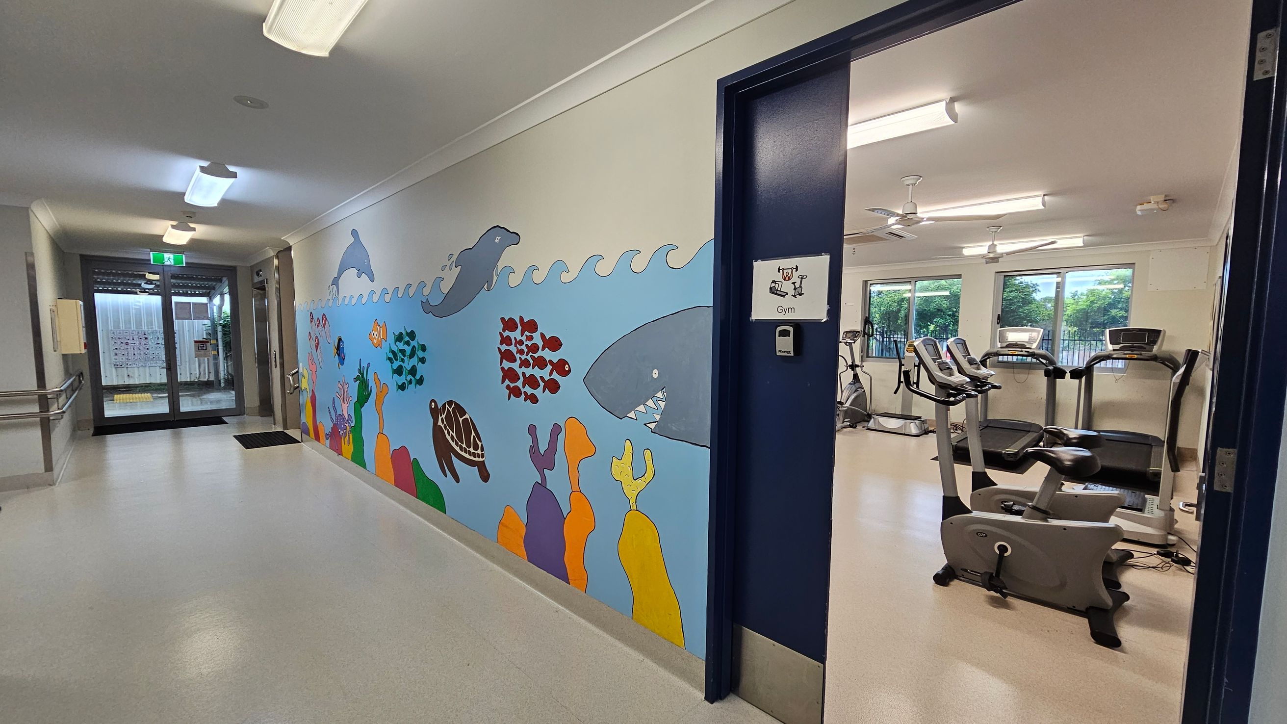 A hallway with a light-coloured floor and ceiling, featuring a colourful mural on the left wall depicting sea creatures like dolphins, fish, a turtle, and a shark. On the right, an open door leads to a gym with exercise equipment, including treadmills and stationary bikes. The gym has large windows with a view of greenery outside. The hallway ends with glass doors leading outside, and there are ceiling lights and a handrail on the left. A hallway with a light-coloured floor and ceiling, featuring a colourful mural on the left wall depicting sea creatures like dolphins, fish, a turtle, and a shark. On the right, an open door leads to a gym with exercise equipment, including treadmills and stationary bikes. The gym has large windows with a view of greenery outside. The hallway ends with glass doors leading outside, and there are ceiling lights and a handrail on the left.