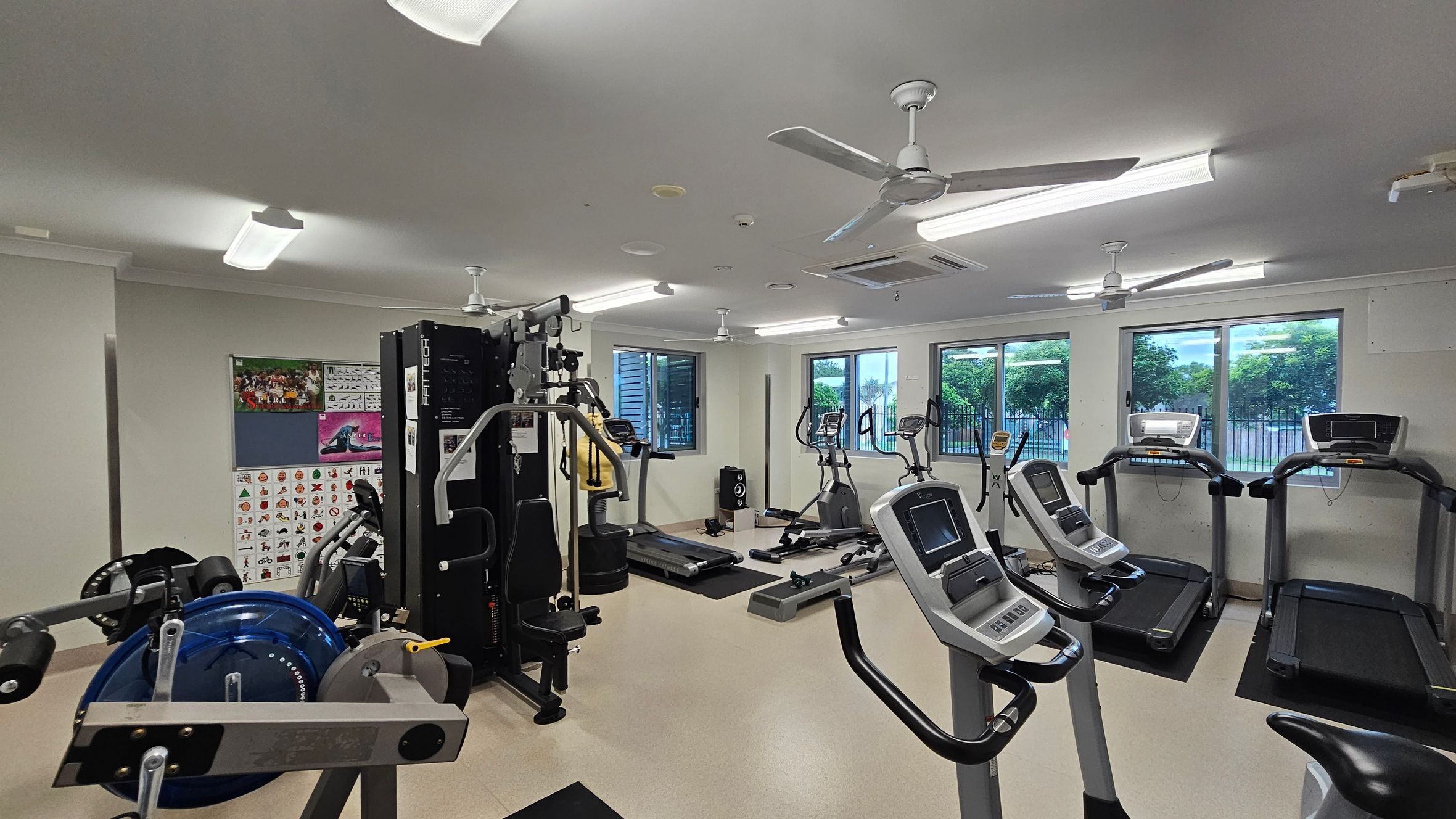 A gym with various exercise equipment. On the left, there is a multi-gym machine and weightlifting equipment. In the middle, there are several exercise bikes and a treadmill. On the right, there are more treadmills. The walls have posters and windows showing greenery outside. The ceiling has fluorescent lights and ceiling fans. The floor is light-coloured and smooth. A gym with various exercise equipment. On the left, there is a multi-gym machine and weightlifting equipment. In the middle, there are several exercise bikes and a treadmill. On the right, there are more treadmills. The walls have posters and windows showing greenery outside. The ceiling has fluorescent lights and ceiling fans. The floor is light-coloured and smooth.