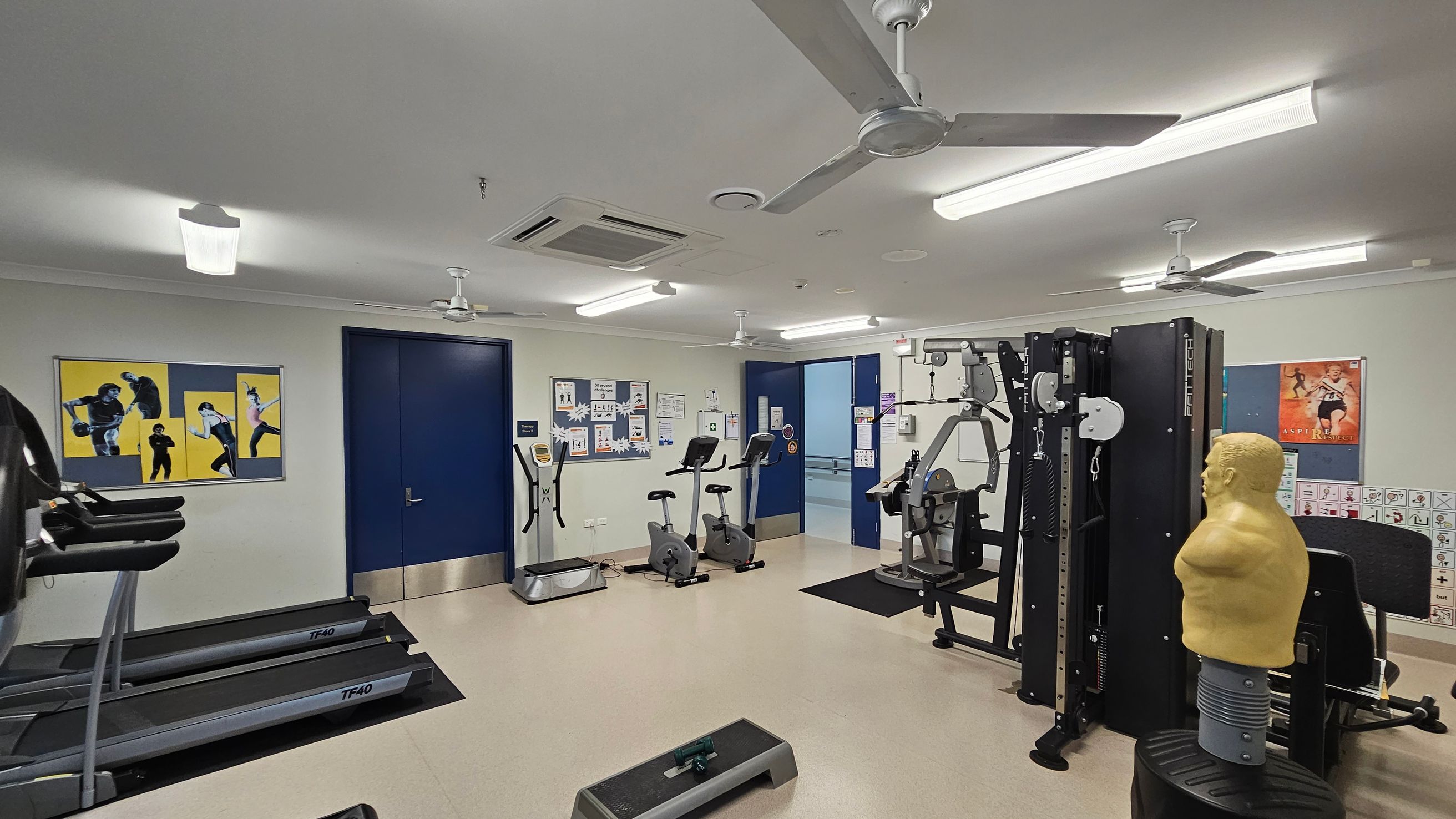 A gym room with various exercise equipment. On the left, there are treadmills and a wall with three fitness posters. In the centre, two stationary bikes and an elliptical machine are positioned near a blue door. The right side features a multi-gym machine and a punching dummy. The walls have motivational posters and informational boards. The ceiling has fluorescent lights and ceiling fans, and the floor is covered with light-coloured tiles. A gym room with various exercise equipment. On the left, there are treadmills and a wall with three fitness posters. In the centre, two stationary bikes and an elliptical machine are positioned near a blue door. The right side features a multi-gym machine and a punching dummy. The walls have motivational posters and informational boards. The ceiling has fluorescent lights and ceiling fans, and the floor is covered with light-coloured tiles.