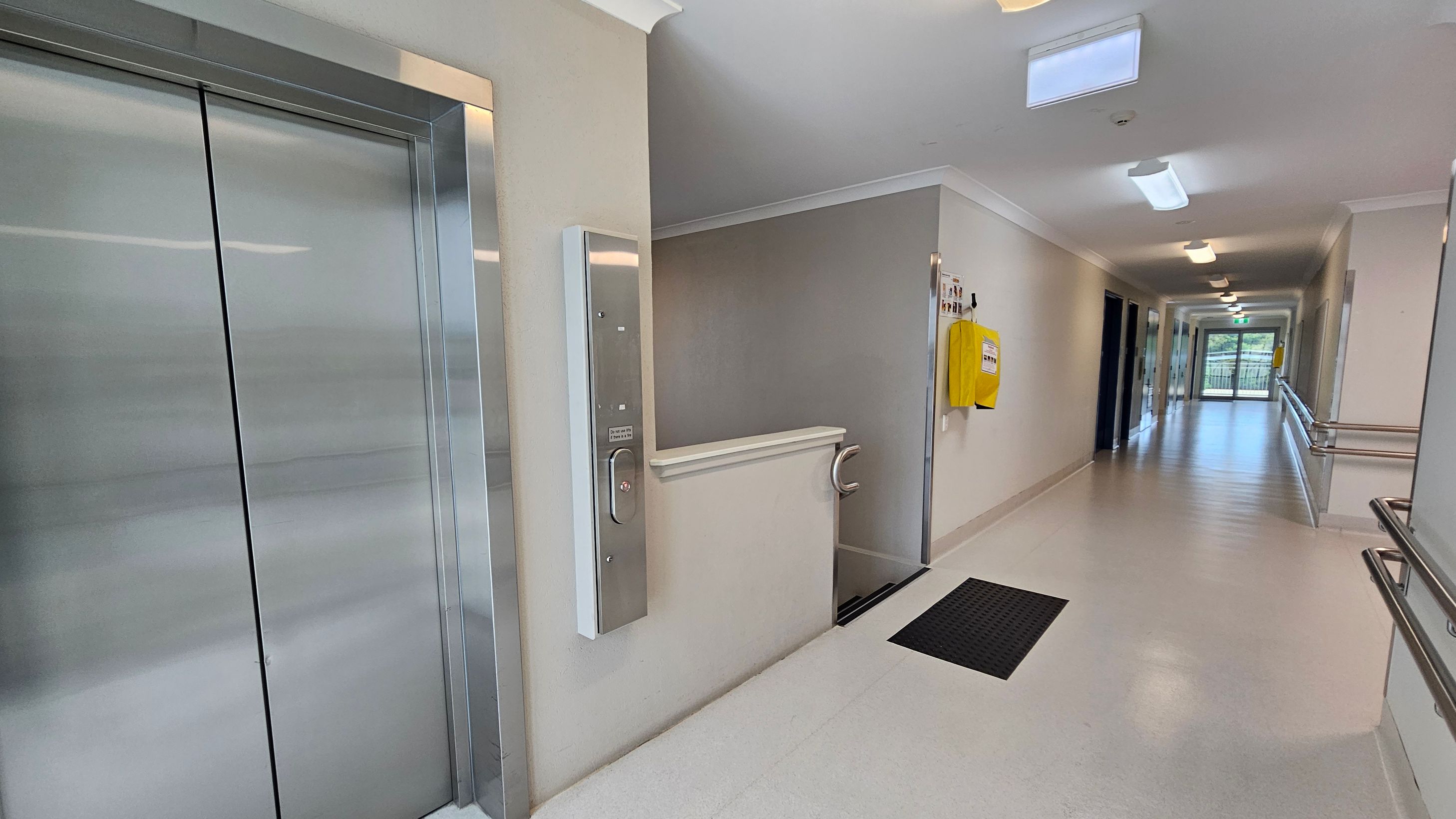 A corridor with a shiny metal lift on the left. Adjacent to the lift is a small platform lift with a control panel. The right side of the corridor features a yellow wall-mounted box and handrails. The floor is light-coloured with a black mat near the platform lift. The ceiling has several rectangular lights, and at the end of the corridor, there is a glass door leading outside. A corridor with a shiny metal lift on the left. Adjacent to the lift is a small platform lift with a control panel. The right side of the corridor features a yellow wall-mounted box and handrails. The floor is light-coloured with a black mat near the platform lift. The ceiling has several rectangular lights, and at the end of the corridor, there is a glass door leading outside.