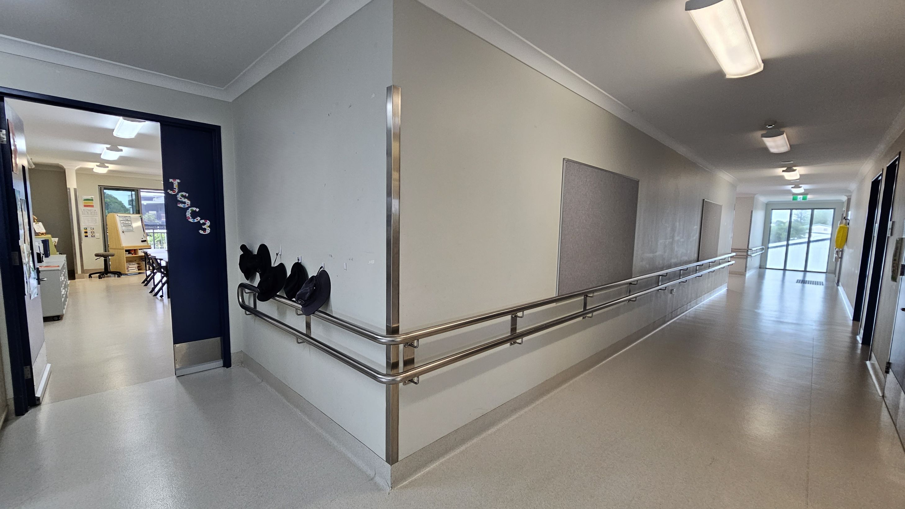 A corridor with a light-coloured floor and ceiling, featuring several overhead lights. On the right, a long handrail runs along the wall, which has a few notice boards. At the end of the corridor, there are glass doors. On the left, an open doorway leads to a classroom with chairs, a desk, and a window. The door has "JSCC3" written on it, and there are hooks with hats hanging on the wall nearby. A corridor with a light-coloured floor and ceiling, featuring several overhead lights. On the right, a long handrail runs along the wall, which has a few notice boards. At the end of the corridor, there are glass doors. On the left, an open doorway leads to a classroom with chairs, a desk, and a window. The door has "JSCC3" written on it, and there are hooks with hats hanging on the wall nearby.