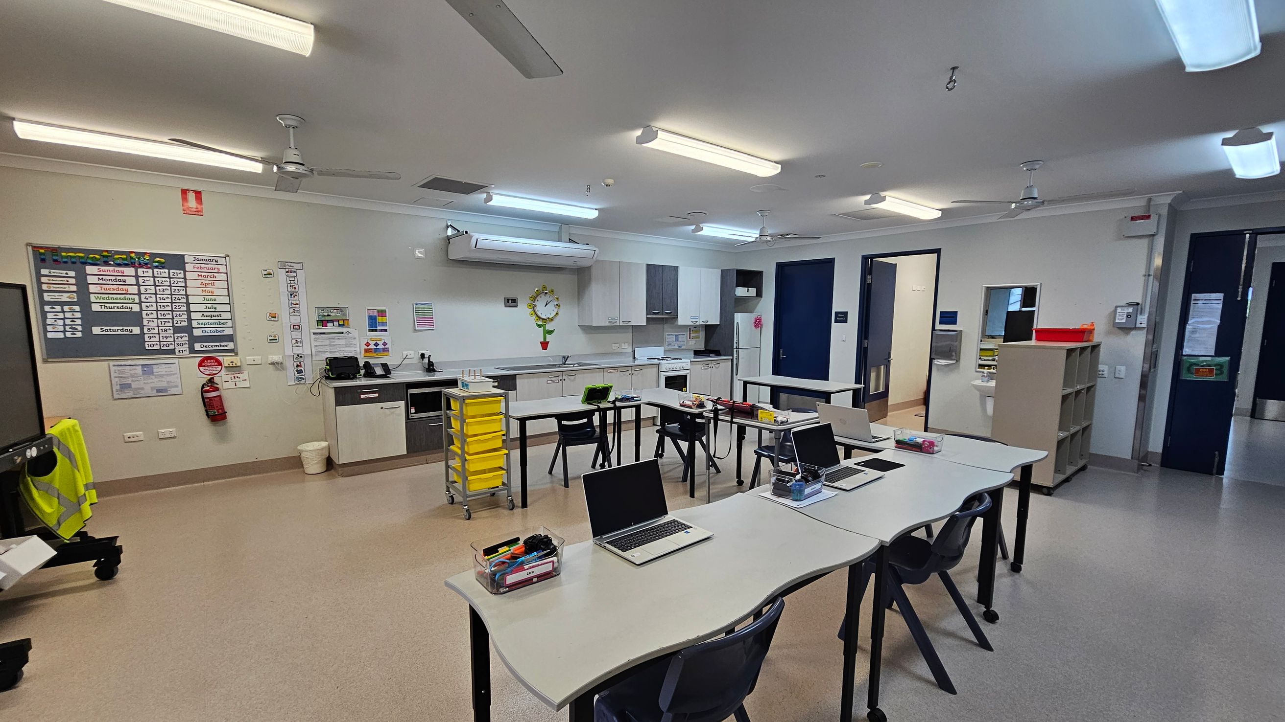 A classroom with a light grey floor and ceiling. In the foreground, there are several white desks with laptops and stationery. The background features a kitchenette with white cabinets, a wall clock, and a yellow storage trolley. On the left wall, there are educational posters and a fire extinguisher. The right side has a doorway and a shelf with a red tray on top. Fluorescent lights and ceiling fans are visible above. A classroom with a light grey floor and ceiling. In the foreground, there are several white desks with laptops and stationery. The background features a kitchenette with white cabinets, a wall clock, and a yellow storage trolley. On the left wall, there are educational posters and a fire extinguisher. The right side has a doorway and a shelf with a red tray on top. Fluorescent lights and ceiling fans are visible above.