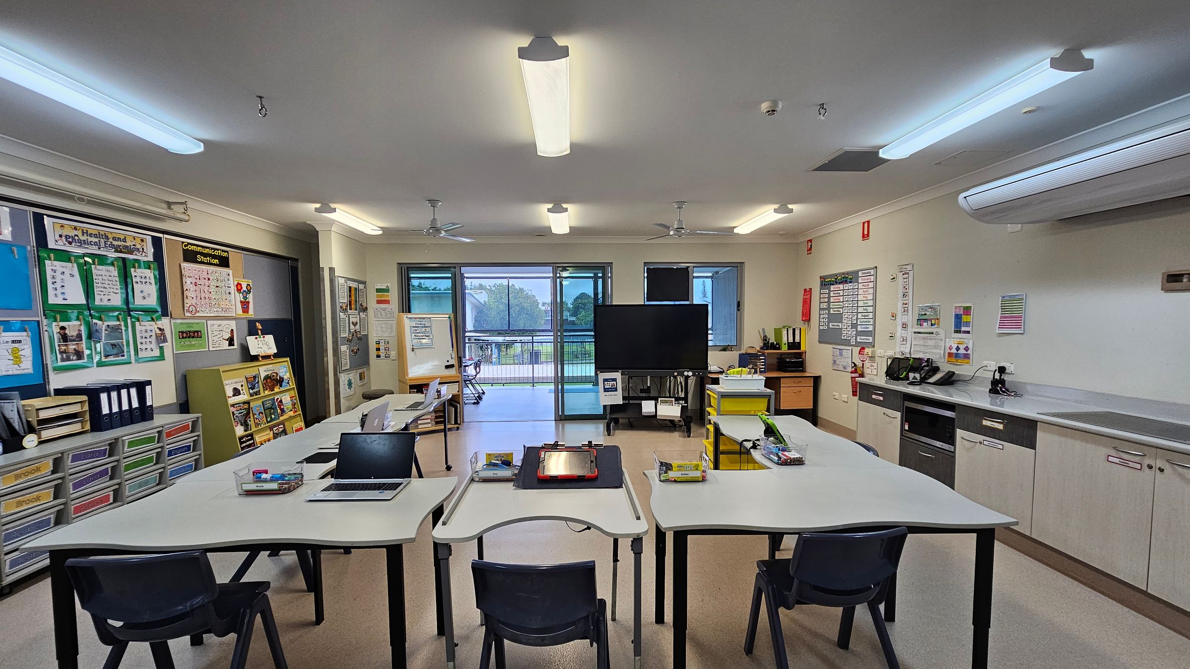 Classroom with a U-shaped arrangement of white tables and black chairs in the foreground. Laptops and stationery are on the tables. The left wall has educational posters and storage drawers. The right side features a counter with cabinets and office equipment. In the centre, a large screen on a stand is near sliding glass doors leading to an outdoor area. The ceiling has fluorescent lights and fans, and the floor is light-coloured. Classroom with a U-shaped arrangement of white tables and black chairs in the foreground. Laptops and stationery are on the tables. The left wall has educational posters and storage drawers. The right side features a counter with cabinets and office equipment. In the centre, a large screen on a stand is near sliding glass doors leading to an outdoor area. The ceiling has fluorescent lights and fans, and the floor is light-coloured.