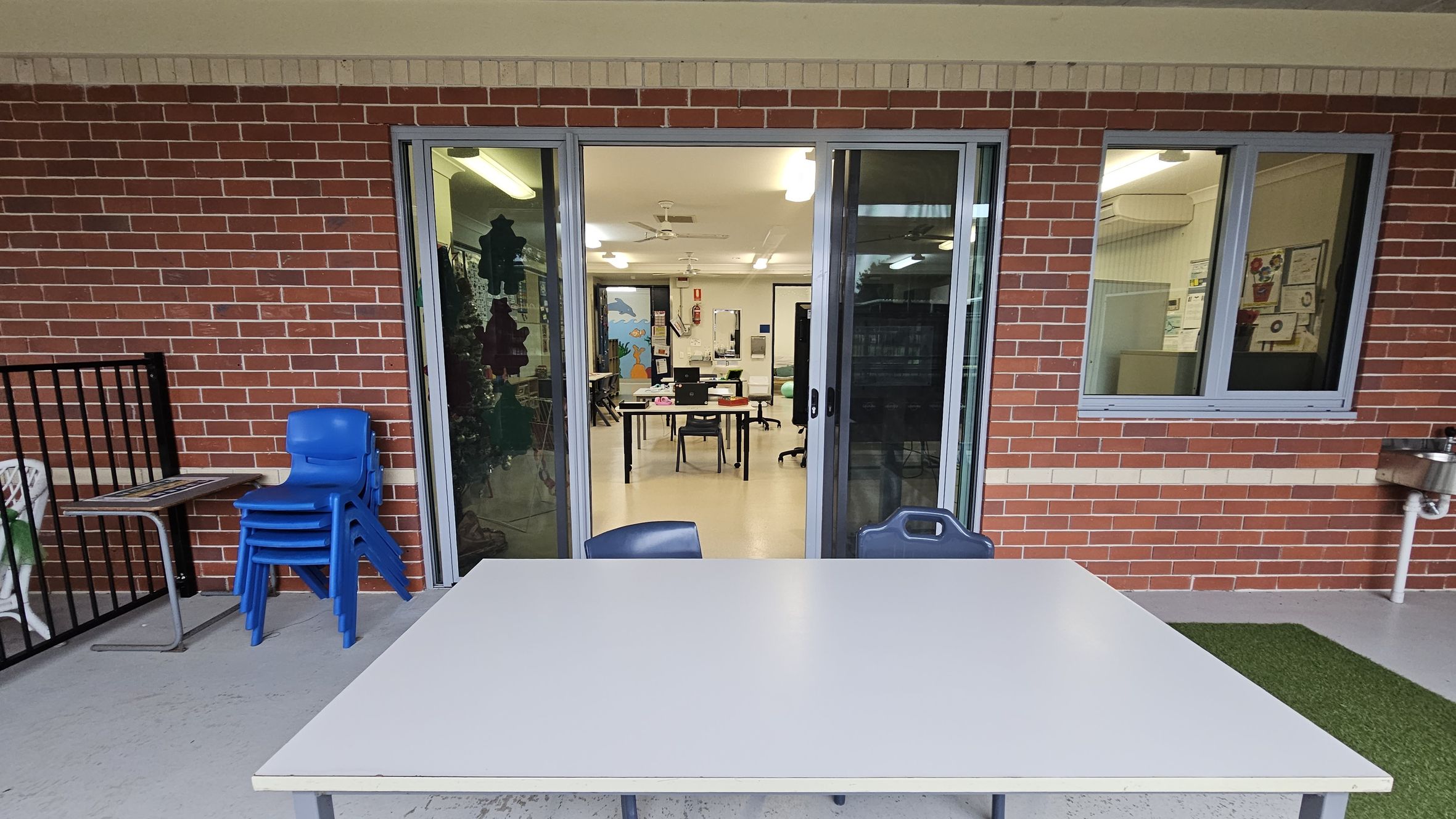 A brick wall with a sliding glass door and a window. In front of the door, there's a white table with a blue chair. To the left, a stack of blue chairs is next to a small table. Inside, a classroom is visible with tables, chairs, and a colourful mural on the far wall. The floor is concrete, and there's a patch of artificial grass on the right. A drinking fountain is mounted on the wall to the right. Ceiling lights are visible inside the room. A brick wall with a sliding glass door and a window. In front of the door, there's a white table with a blue chair. To the left, a stack of blue chairs is next to a small table. Inside, a classroom is visible with tables, chairs, and a colourful mural on the far wall. The floor is concrete, and there's a patch of artificial grass on the right. A drinking fountain is mounted on the wall to the right. Ceiling lights are visible inside the room.