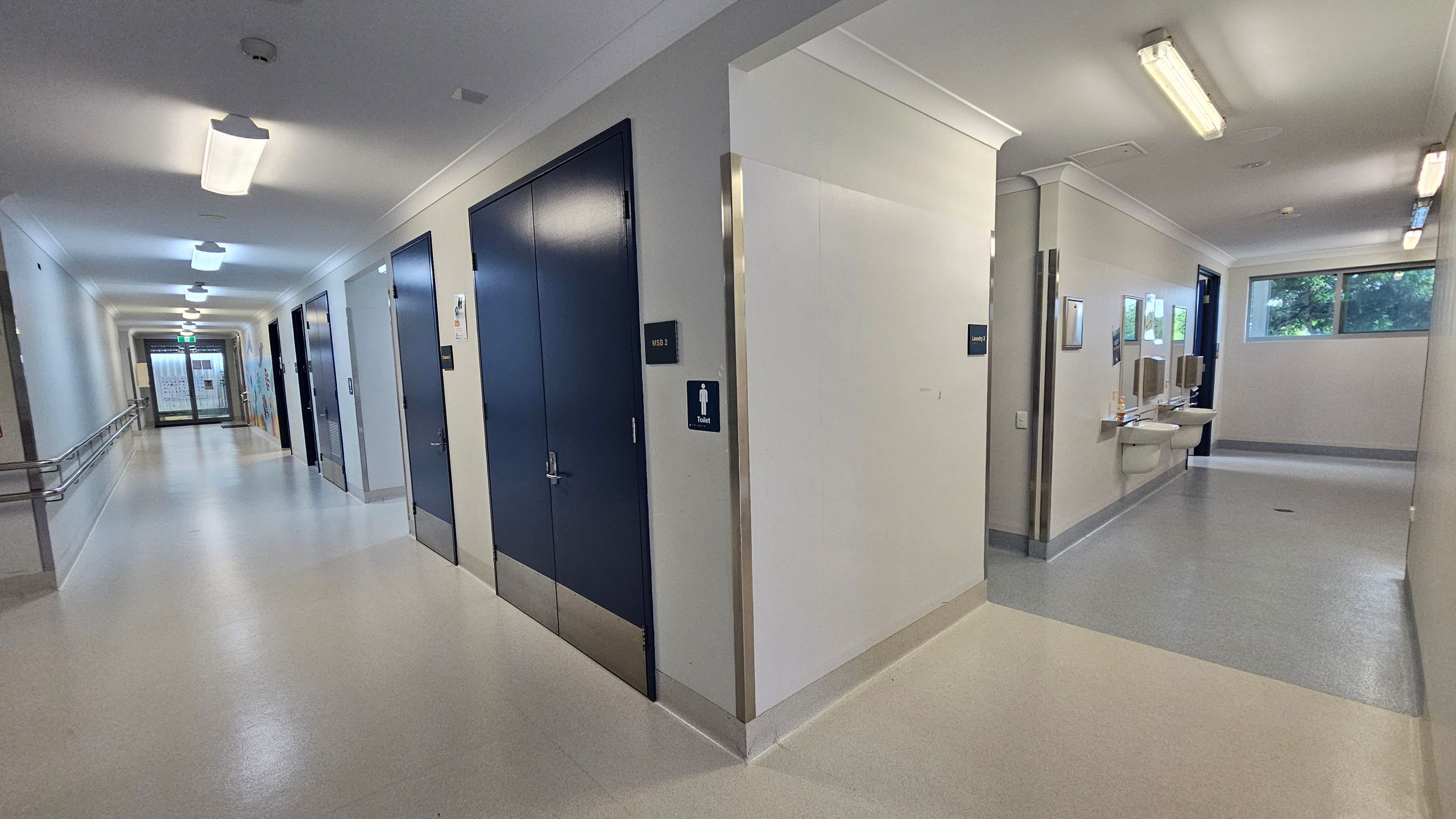 A corridor with a light-coloured floor and ceiling. On the left, there are several doors with signs, and a handrail runs along the wall. On the right, a section leads to a toilet area with sinks and mirrors. Fluorescent lights are mounted on the ceiling. At the end of the corridor, there is a glass door with a view of a window showing greenery outside. A corridor with a light-coloured floor and ceiling. On the left, there are several doors with signs, and a handrail runs along the wall. On the right, a section leads to a toilet area with sinks and mirrors. Fluorescent lights are mounted on the ceiling. At the end of the corridor, there is a glass door with a view of a window showing greenery outside.