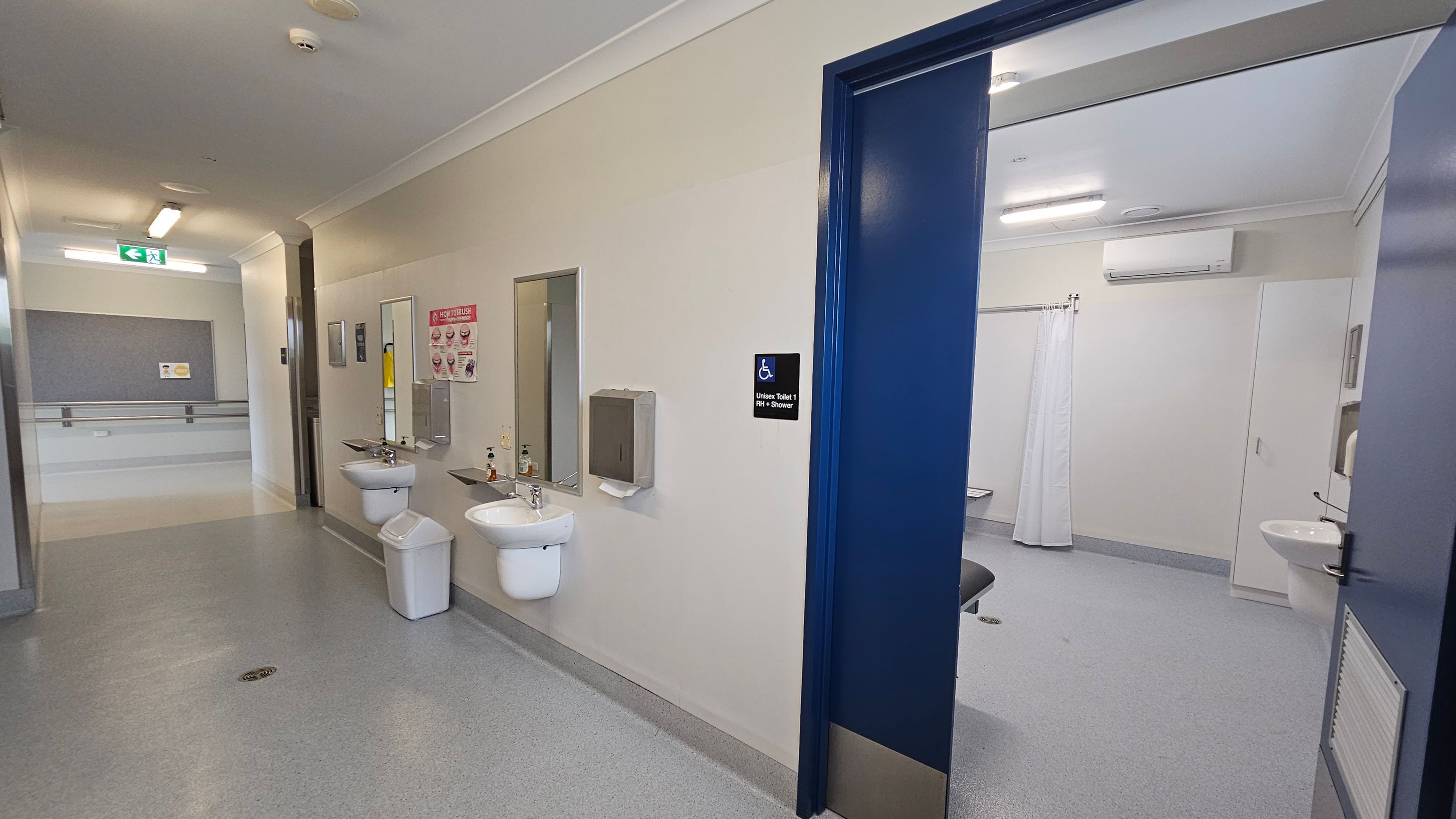 A corridor with a grey floor and white ceiling. On the left, two sinks with mirrors and soap dispensers are mounted on the wall, along with a paper towel dispenser. A sign indicates a unisex toilet with a shower. The right side shows an open door leading to a bathroom with a white curtain and a sink. An air conditioning unit is visible above. The corridor has a handrail and an emergency exit sign. A corridor with a grey floor and white ceiling. On the left, two sinks with mirrors and soap dispensers are mounted on the wall, along with a paper towel dispenser. A sign indicates a unisex toilet with a shower. The right side shows an open door leading to a bathroom with a white curtain and a sink. An air conditioning unit is visible above. The corridor has a handrail and an emergency exit sign.