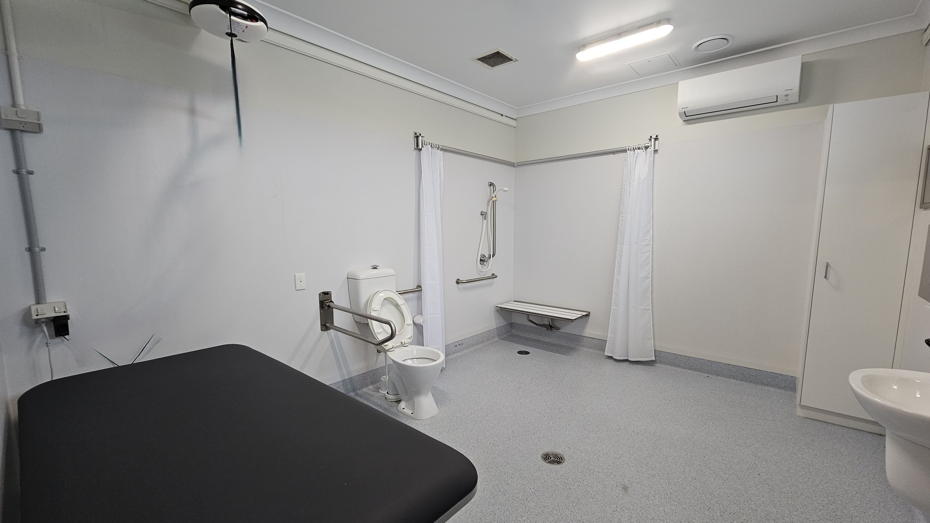 A spacious, accessible bathroom with a grey floor and white walls. On the left, a black padded bench is visible. In the centre, a toilet with a support rail is next to a shower area with a foldable seat and a curtain. On the right, a white sink is partially visible, and an air conditioning unit is mounted above a closed cabinet. The ceiling is white with a fluorescent light and a vent. A spacious, accessible bathroom with a grey floor and white walls. On the left, a black padded bench is visible. In the centre, a toilet with a support rail is next to a shower area with a foldable seat and a curtain. On the right, a white sink is partially visible, and an air conditioning unit is mounted above a closed cabinet. The ceiling is white with a fluorescent light and a vent.