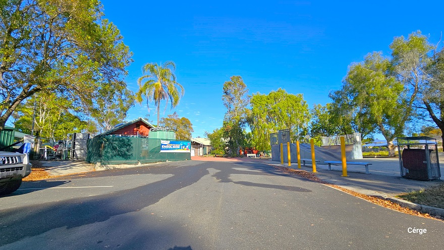 Entryway from the carpark leading to the entrance of Dayboro Pool & Gym. Tall yellow bollards marked the boundary of the skate park on the right side. A large grey metal garbage bin with a canopy cover is placed before two benches at the back of the bollards. Covering the perimeter of the facility is a green cloth on the left, while on the right is white. Tall trees with great foliage offer shade to the surrounding area. Entryway from the carpark leading to the entrance of Dayboro Pool & Gym. Tall yellow bollards marked the boundary of the skate park on the right side. A large grey metal garbage bin with a canopy cover is placed before two benches at the back of the bollards. Covering the perimeter of the facility is a green cloth on the left, while on the right is white. Tall trees with great foliage offer shade to the surrounding area.