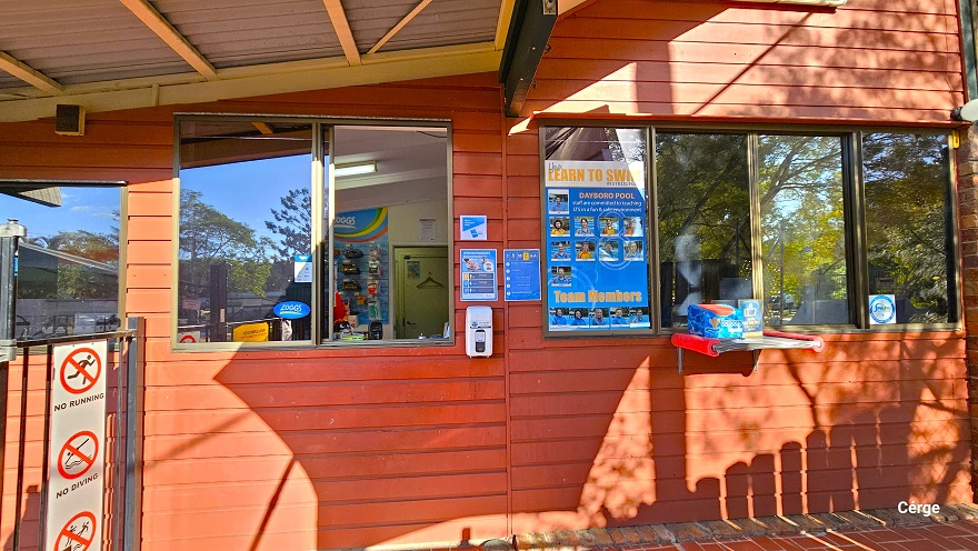 Entry to Dayboro Pool & Gym is through a black chain-link gate with grey metal tube frames and a red brick floor. Guest admittance is to the glass windows of the small, low-set 1970s-style red brick building with a corrugated iron roof. In front of it is a low black fence and a purple sunshade. Behind the left gate are plants, while behind the right gate are ‘Smoke-free healthcare” and “NO Dogs allowed on premises' ' signs placed on the brick wall. Entry to Dayboro Pool & Gym is through a black chain-link gate with grey metal tube frames and a red brick floor. Guest admittance is to the glass windows of the small, low-set 1970s-style red brick building with a corrugated iron roof. In front of it is a low black fence and a purple sunshade. Behind the left gate are plants, while behind the right gate are ‘Smoke-free healthcare” and “NO Dogs allowed on premises' ' signs placed on the brick wall.