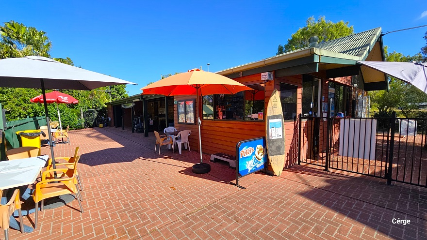 The outdoor seating area in Dayboro Pool & Gym has a natural vibe setting with an overlook of the tall trees with abundant foliage. There are glass windows on this side of the building, and it has a similar red brick floor to the reception. The white, orange, and red sun shades have orange, white plastic chairs, and a white table. A tall yellow garbage bin with a black garbage bag is placed against the green curvy-style fence. A wooden surfing board is left against the wall near the black fence. The outdoor seating area in Dayboro Pool & Gym has a natural vibe setting with an overlook of the tall trees with abundant foliage. There are glass windows on this side of the building, and it has a similar red brick floor to the reception. The white, orange, and red sun shades have orange, white plastic chairs, and a white table. A tall yellow garbage bin with a black garbage bag is placed against the green curvy-style fence. A wooden surfing board is left against the wall near the black fence.