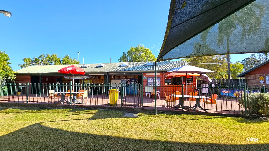 View of the seating area in front of the low-set 1970s-style red brick building when standing inside the green curvy-style fence. Within the green fence is an open area with a well-maintained height. This image also shows the corrugated roof of the building with square bumps. View of the seating area in front of the low-set 1970s-style red brick building when standing inside the green curvy-style fence. Within the green fence is an open area with a well-maintained height. This image also shows the corrugated roof of the building with square bumps.