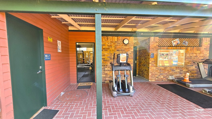 The 1970-style brick building with red corrugated walls and roofing has a red brick floor style. The building has green metal frames as support. Within the red corrugated wall is the accessible bathroom, which can be distinguished by the green door, a black mat, and a blue Unisex Toilet label. On the brick wall to the right is a painting and two photos. Two elliptical trainers are available in this area. The 1970-style brick building with red corrugated walls and roofing has a red brick floor style. The building has green metal frames as support. Within the red corrugated wall is the accessible bathroom, which can be distinguished by the green door, a black mat, and a blue Unisex Toilet label. On the brick wall to the right is a painting and two photos. Two elliptical trainers are available in this area.