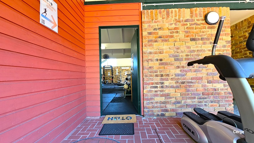 Entryway to Dayboro Gym. The gym can be identified by the red-to-orange brick wall and red corrugated wall and a directional white label placed within this wall that details the Dayboro Gym with an arrow pointing to the door. There is a brown floor mat with a Hello detail on it and a black mat for dirt removal before entering the amenity. Found on the right is cardio equipment called an Elliptical trainer. Entryway to Dayboro Gym. The gym can be identified by the red-to-orange brick wall and red corrugated wall and a directional white label placed within this wall that details the Dayboro Gym with an arrow pointing to the door. There is a brown floor mat with a Hello detail on it and a black mat for dirt removal before entering the amenity. Found on the right is cardio equipment called an Elliptical trainer.
