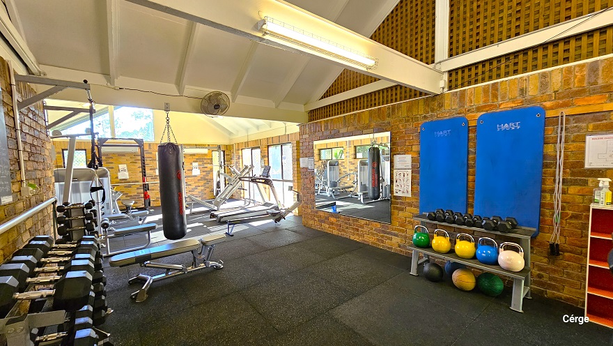 Inside the Dayboro Gym are black dumbbells, a row of colorful kettlebells on a rack, and other cardio endurance equipment. The room has a wooden netted-like design on the upper right wall that has white metal frames. Behind the rack for dumbbells and kettlebells are blue exercise mats and grey ropes. Next to it is a mirror that reflects other gym equipment inside. Further to the back, on the right wall, are glass windows that allow natural light to filter through, illuminating the area inside. Inside the Dayboro Gym are black dumbbells, a row of colorful kettlebells on a rack, and other cardio endurance equipment. The room has a wooden netted-like design on the upper right wall that has white metal frames. Behind the rack for dumbbells and kettlebells are blue exercise mats and grey ropes. Next to it is a mirror that reflects other gym equipment inside. Further to the back, on the right wall, are glass windows that allow natural light to filter through, illuminating the area inside.
