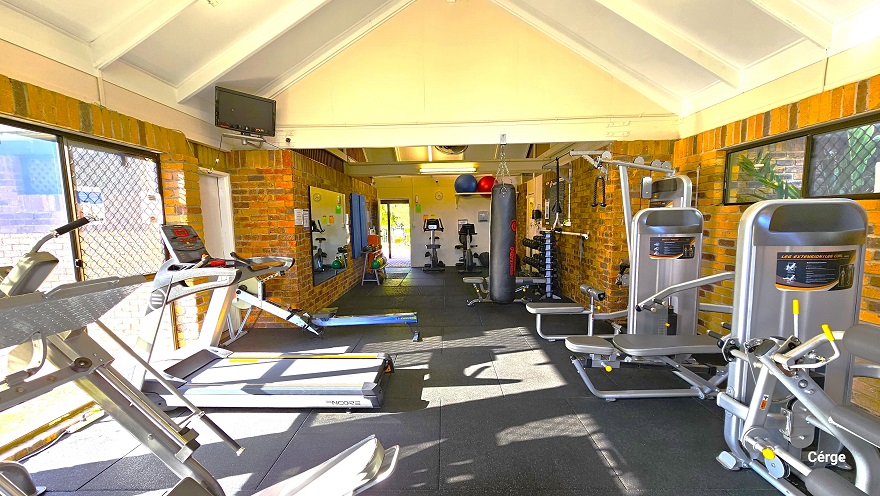 The image shows the back portion of the inside of Dayboro Gym. Mounted on the left wall is a small television above another white door. The glass windows reflect the silhouette of the chain-linked fence outside the building. On the upper left of the white wall next to the entrance door, there are blue and red yoga balls placed on a white rectangular frame. Two small glass windows can be found on the upper part of the right brick wall. The image shows the back portion of the inside of Dayboro Gym. Mounted on the left wall is a small television above another white door. The glass windows reflect the silhouette of the chain-linked fence outside the building. On the upper left of the white wall next to the entrance door, there are blue and red yoga balls placed on a white rectangular frame. Two small glass windows can be found on the upper part of the right brick wall.