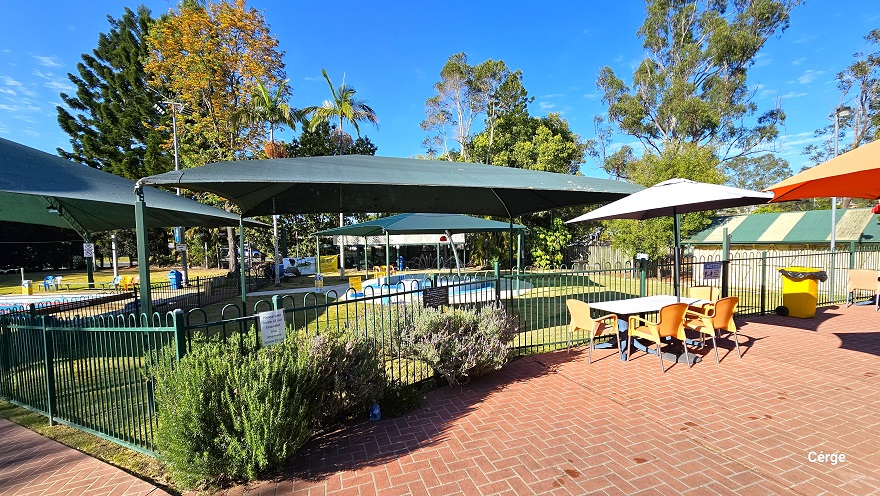 Red brick pathway leading to the six-lane, 25m lap outdoor pool. The pool is situated down a slight incline with a green sun shade on one end of the pool. A yellow sign board can be found under the green shade. To the left is a shed with a green corrugated wall and opening. Two green garbage bins with wheels are placed in front of the shed. To the right is another pool enclosed within a low green pool fence and is covered by two green sun shades. Red brick pathway leading to the six-lane, 25m lap outdoor pool. The pool is situated down a slight incline with a green sun shade on one end of the pool. A yellow sign board can be found under the green shade. To the left is a shed with a green corrugated wall and opening. Two green garbage bins with wheels are placed in front of the shed. To the right is another pool enclosed within a low green pool fence and is covered by two green sun shades.