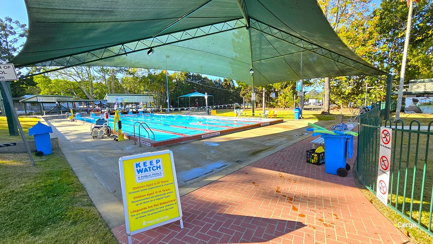 Closer of the six-lane, 25m lap outdoor pool. A yellow sign board that reminds adults to keep watch for children of different age brackets and what lifeguards do can be found on the edge of the red brick pathway that extends to the right. Within this pathway is a grey metal bench, and a blue container with black wheels that contains lime green and blue plastic tubes. There is an aquatic pool and a yellow pool hoist on the left pool deck. Closer of the six-lane, 25m lap outdoor pool. A yellow sign board that reminds adults to keep watch for children of different age brackets and what lifeguards do can be found on the edge of the red brick pathway that extends to the right. Within this pathway is a grey metal bench, and a blue container with black wheels that contains lime green and blue plastic tubes. There is an aquatic pool and a yellow pool hoist on the left pool deck.