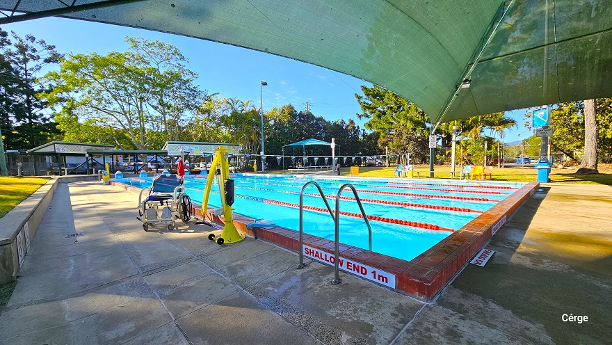 A 25m, 6-lane outdoor pool at Dayboro Pool and Gym with partial shading on one end. The pool has red-tiled edges all around with stainless steel pool ladders. A ‘Shallow end 1m’ and ‘No smoking’ sign is visible on the edge of the pool. There is an accessible aquatic wheelchair and a yellow pool hoist on the left pool deck. Each lane is separated by blue, red, and white floating rope lanes. Overhang the pool is strings of grey triangular flags wrapped around a pole on the opposite pool sides. A 25m, 6-lane outdoor pool at Dayboro Pool and Gym with partial shading on one end. The pool has red-tiled edges all around with stainless steel pool ladders. A ‘Shallow end 1m’ and ‘No smoking’ sign is visible on the edge of the pool. There is an accessible aquatic wheelchair and a yellow pool hoist on the left pool deck. Each lane is separated by blue, red, and white floating rope lanes. Overhang the pool is strings of grey triangular flags wrapped around a pole on the opposite pool sides.
