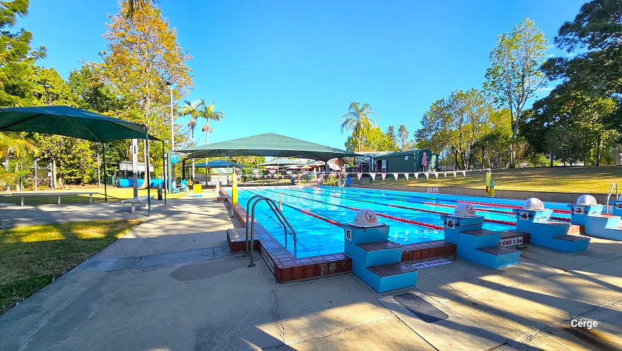 View from the diving platform of the 25m, 6-lane outdoor pool at Dayboro Pool and Gym. The pool deck has a rough surface floor to help avoid slipping. Each lane has a two-step stair with a grey non-slip cover blue diving platform. On top of the diving platform is a yellow semi-circle with a red caution on it. There is a green sunshade on the left with grey metal benches. Yellow floating devices are wrapped around poles with strings of blue triangular flags. View from the diving platform of the 25m, 6-lane outdoor pool at Dayboro Pool and Gym. The pool deck has a rough surface floor to help avoid slipping. Each lane has a two-step stair with a grey non-slip cover blue diving platform. On top of the diving platform is a yellow semi-circle with a red caution on it. There is a green sunshade on the left with grey metal benches. Yellow floating devices are wrapped around poles with strings of blue triangular flags.