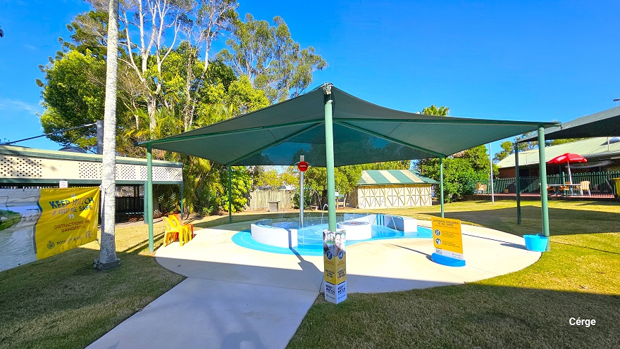 There is a smooth concrete pathway leading to the small toddler splash pool at Dayboro Pool & Gym. The pool is protected by a green sunshade wrapped around green posts. On a silver metal post is a large red lip graphic placed near the top of the post. The splash toddler aquatic activity offers interactive narrow water fountains from the two opposite white curves. There are yellow and red monoblock plastic chairs on the pool deck. There is a smooth concrete pathway leading to the small toddler splash pool at Dayboro Pool & Gym. The pool is protected by a green sunshade wrapped around green posts. On a silver metal post is a large red lip graphic placed near the top of the post. The splash toddler aquatic activity offers interactive narrow water fountains from the two opposite white curves. There are yellow and red monoblock plastic chairs on the pool deck.