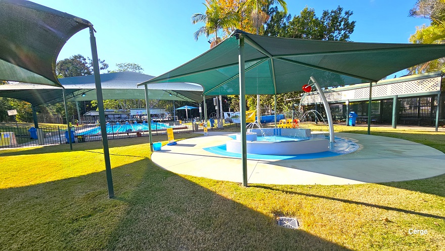 The image shows the side of the small toddler splash pool. The red lip graphic is attached to a red semi-circle on a curvy silver post. The splash toddler aquatic activity offers interactive narrow water fountains from the two opposite white curves. There are yellow and red monoblock plastic chairs on the pool deck. Further to the right is the long sunshade with an upper-side white cover that has several holes. At the back is the 25m, 6-lane outdoor pool. The image shows the side of the small toddler splash pool. The red lip graphic is attached to a red semi-circle on a curvy silver post. The splash toddler aquatic activity offers interactive narrow water fountains from the two opposite white curves. There are yellow and red monoblock plastic chairs on the pool deck. Further to the right is the long sunshade with an upper-side white cover that has several holes. At the back is the 25m, 6-lane outdoor pool.