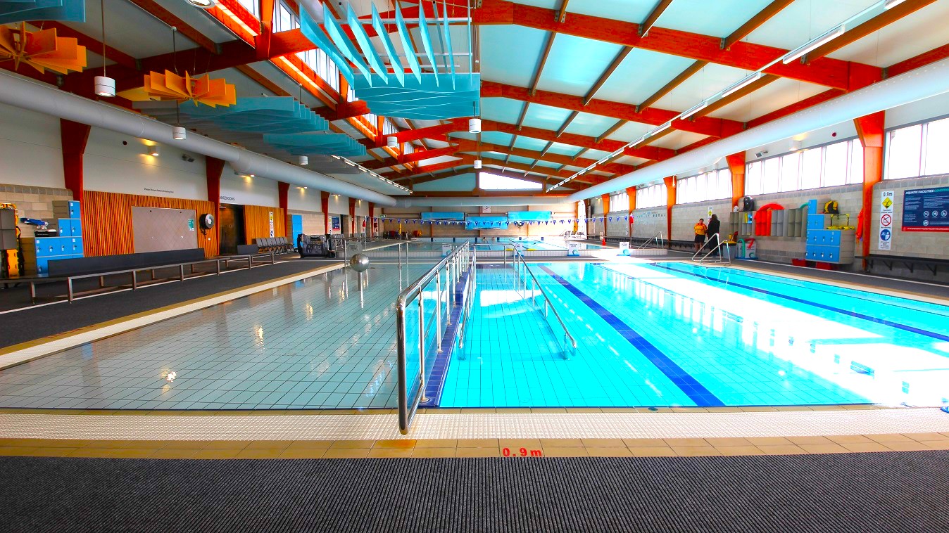 SWIRL Aquatic and Recreation Centre is a low-set warehouse-style facility with features painted in white and light green and a range of small trees and shrubs surrounding it. A grey fence secures the perimeter. Turning right from Nelson St and down the sloping driveway is the dedicated parking space with two accessible car parks.