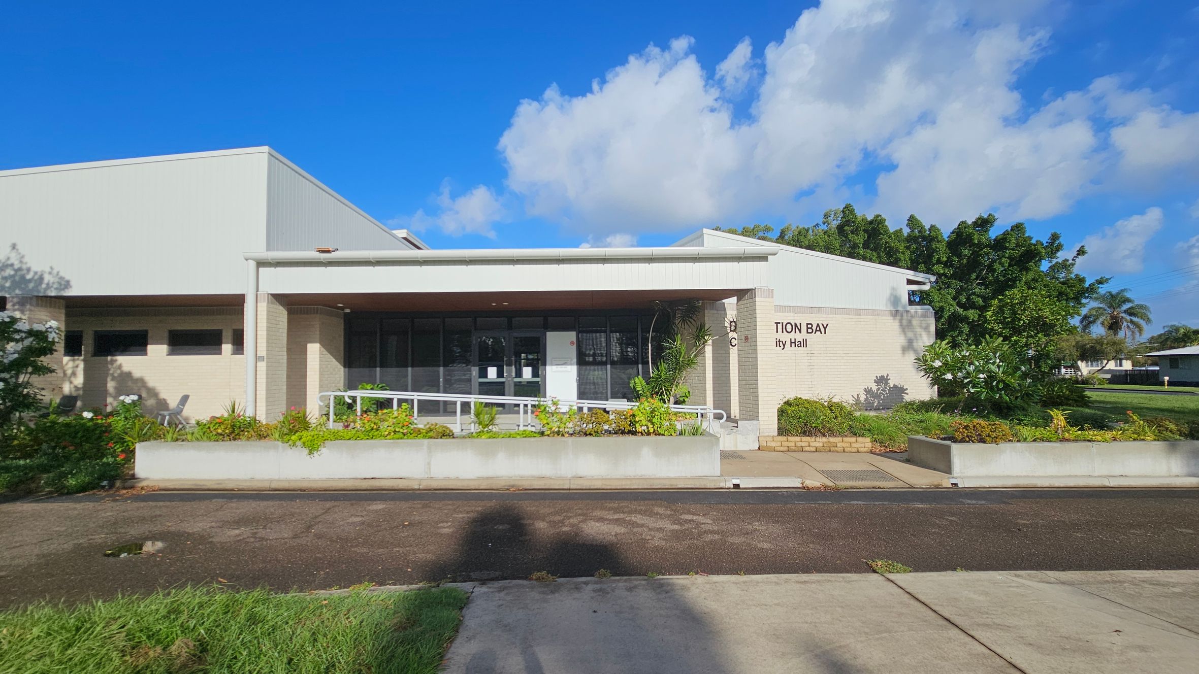 A single-storey building with a flat roof and light-coloured brick exterior. The entrance features large glass windows and a ramp with a white railing. In front, there are rectangular planters with various green plants and shrubs. The building's name is partially visible on the right side. The foreground has a paved path and grass, while the background includes trees and a clear blue sky with some clouds.