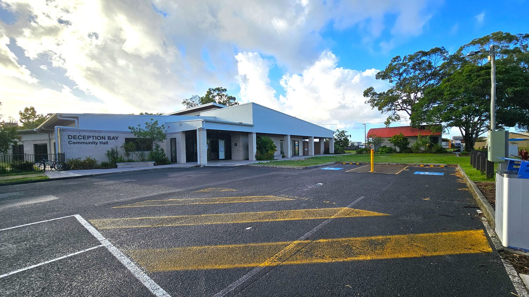 A community hall with a white exterior and a sign reading "Deception Bay Community Hall" on the left. The building has a modern design with a sloped roof and large windows. In front, there is a paved parking area with yellow markings and a few designated parking spaces. On the right, there are large trees and a smaller building with a red roof. The sky is partly cloudy, and the area is surrounded by greenery.