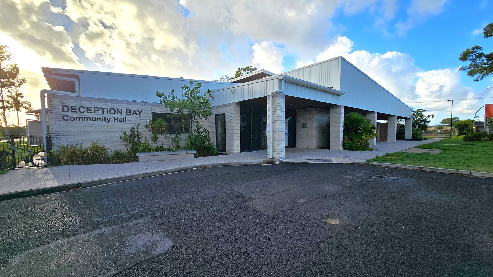 A modern building with a white exterior and a flat roof, labelled "Deception Bay Community Hall" on the left side. The entrance features glass doors and is surrounded by small plants and shrubs. A bicycle is parked near the entrance. The foreground shows a paved road, while the background includes a grassy area with trees and a clear sky. The building has a covered walkway supported by white pillars.