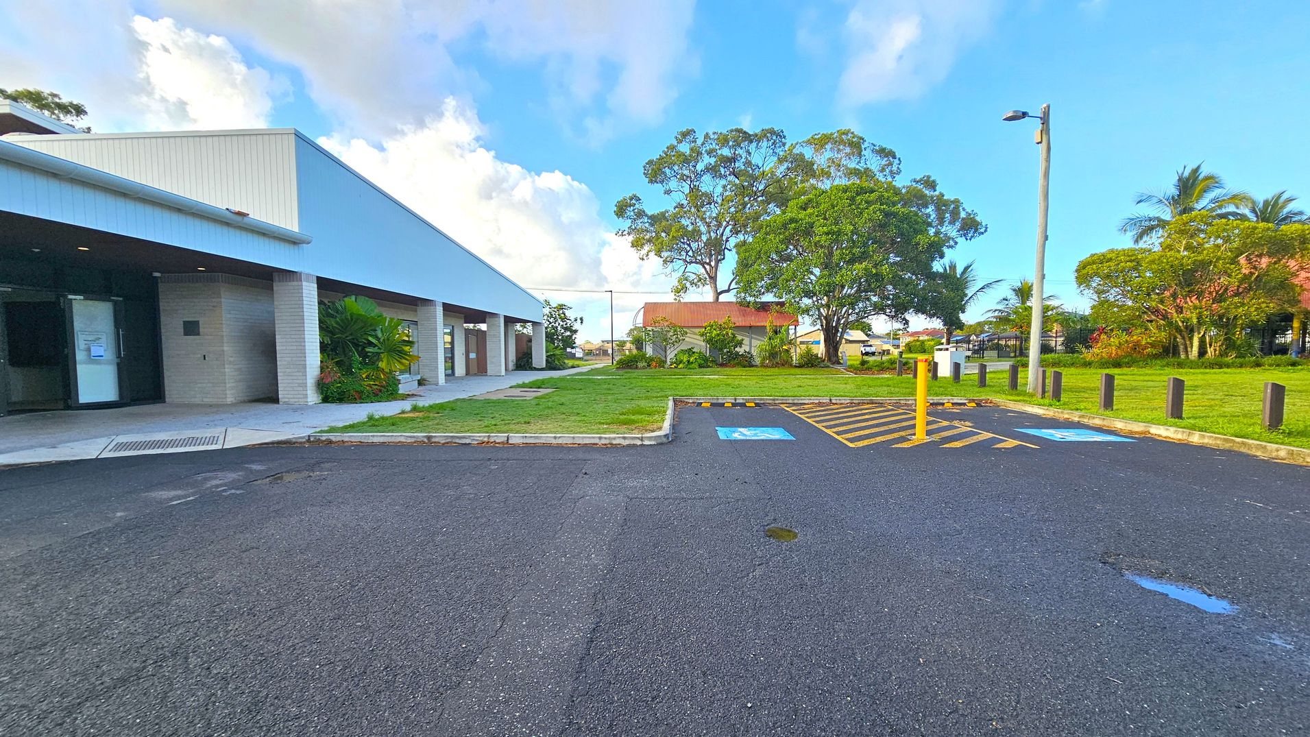 A building with a sloped roof and brick columns is on the left, with a pathway and some plants near the entrance. In the centre, there's a grassy area with a large tree and a smaller building with a rust-coloured roof. On the right, a car park with marked spaces, including a disabled parking spot, is visible. A lamp post and several trees are in the background. The ground is paved, and the sky is partly cloudy.