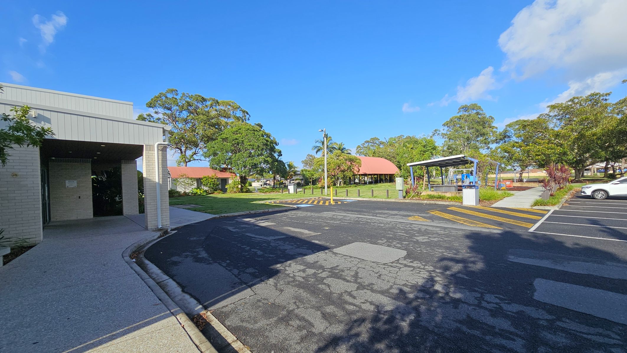 On the left, a light-coloured brick building with a covered entrance and a concrete path. In the centre, a paved road with yellow pedestrian crossing lines leads to a grassy area with trees and a structure with a red roof. On the right, a small shelter with a metal roof and a nearby car park with a white car. The background features more trees and a clear blue sky. Shadows of trees are cast on the road.