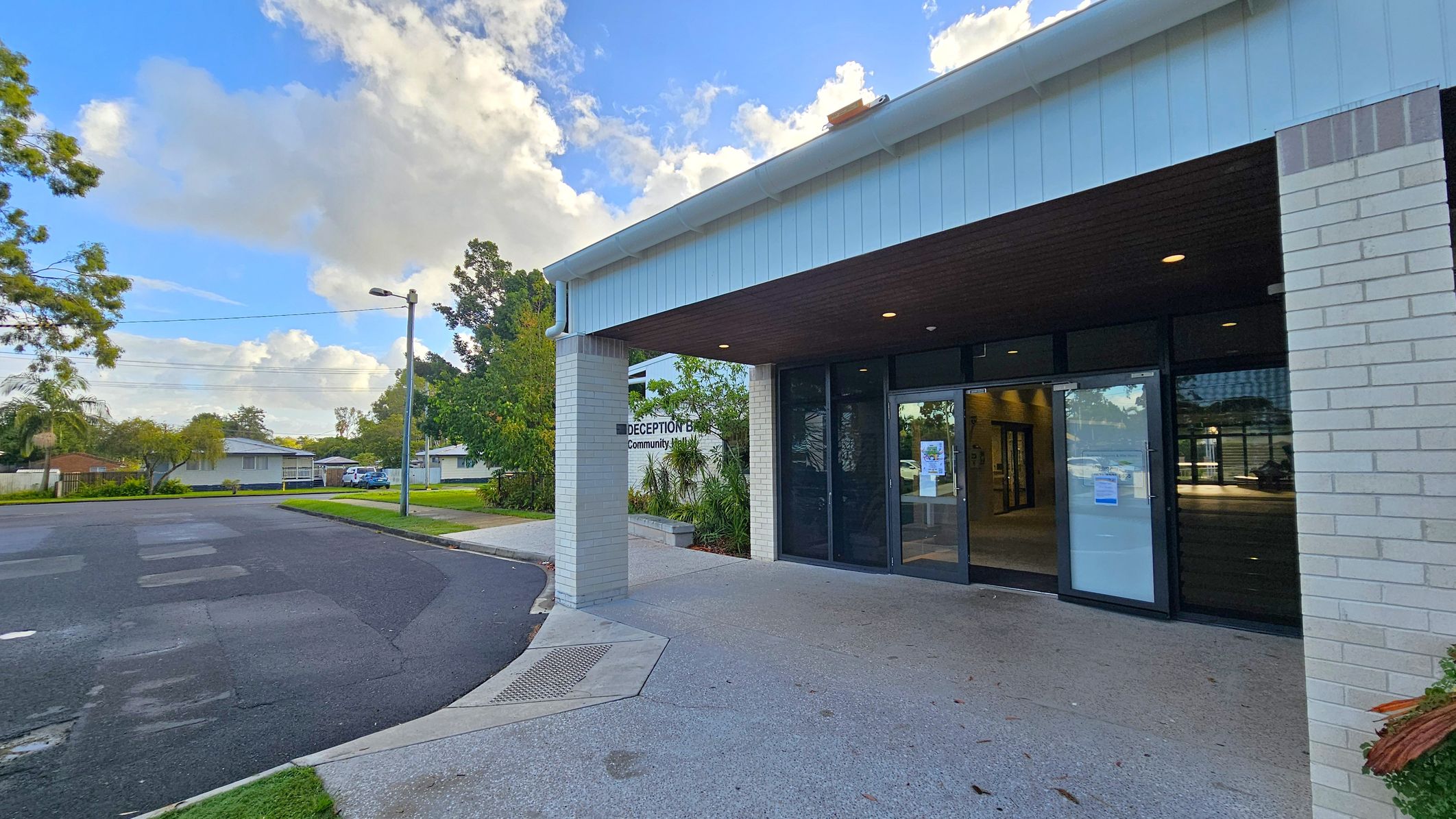 A building entrance with a sign reading "Deception Bay Community Hall" on the right. The structure has a white brick exterior and a dark overhang with recessed lighting. Glass doors lead inside. On the left, a street curves around a grassy area with trees and a few houses in the background. A streetlamp is visible near the road. The pavement in front of the entrance is light grey with a textured surface.
