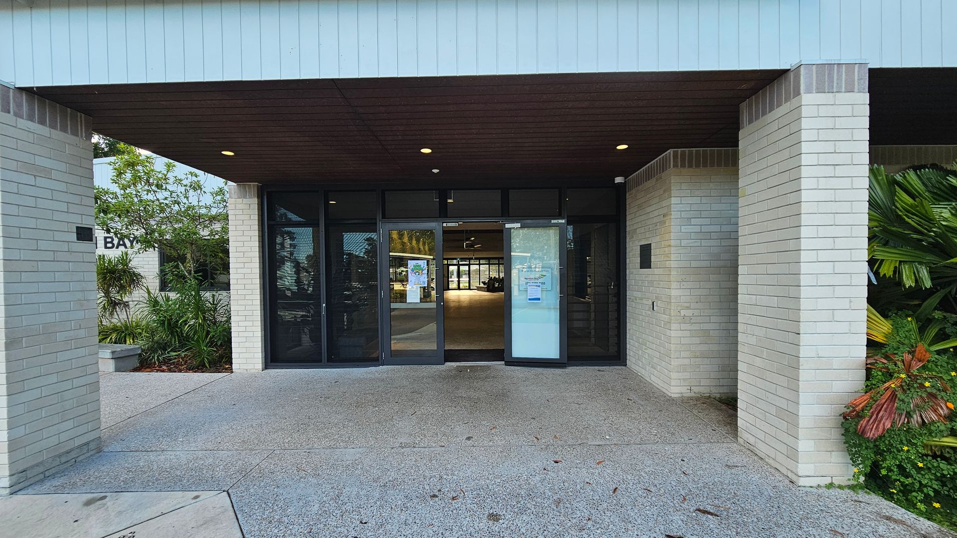 Entrance to a building with light-coloured brick walls and a dark overhang ceiling. The double glass doors are in the centre, with notices posted on them. On the left, there are green plants and a small sign with the word "BAY" partially visible. On the right, more lush greenery is present. The ground is paved with a textured surface.