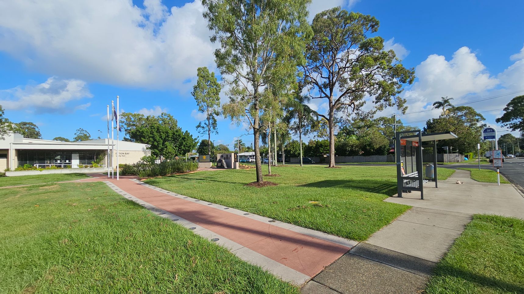 A grassy area with a paved path runs through the centre, flanked by trees. On the left, a building with large windows and flagpoles is visible. On the right, a bus stop with a shelter and bench is situated next to a road. The background features more trees and a sign near the road. The sky is partly cloudy.