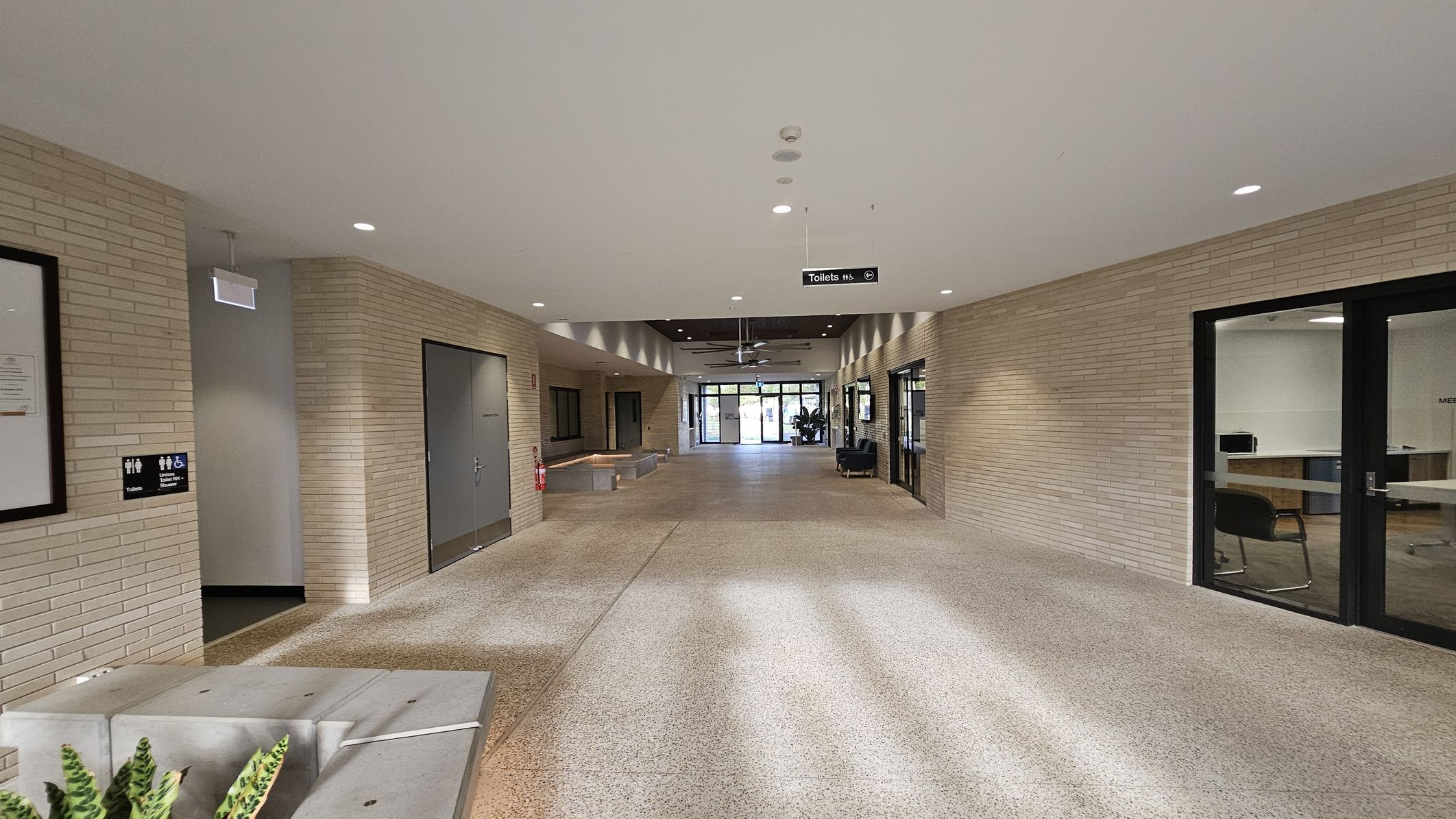 A hallway with light brick walls and a speckled floor. On the left, there are signs for toilets and a framed notice. A grey door is visible further down. On the right, there are glass doors leading to an office space with chairs and a desk. In the centre, a ceiling fan is visible, and at the end of the hallway, there are glass doors leading outside. A small plant is in the foreground on the left. The ceiling is white with recessed lighting.