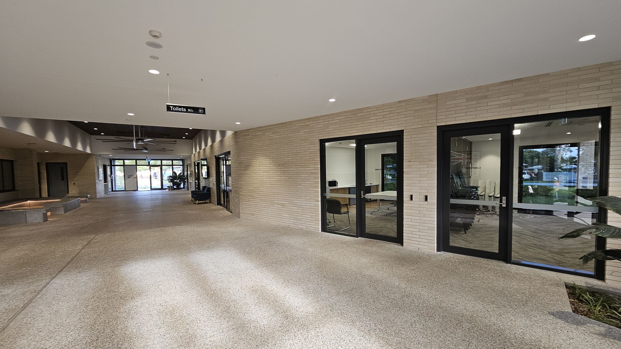 A spacious indoor corridor with a light-coloured, textured floor and a ceiling with recessed lighting. On the right, there are glass doors leading to meeting rooms, with visible tables and chairs inside. The left side features a brick wall with a recessed area and a bench. A sign hanging from the ceiling indicates the direction to the toilets. At the far end, large glass doors open to an outdoor area with greenery visible through the glass.