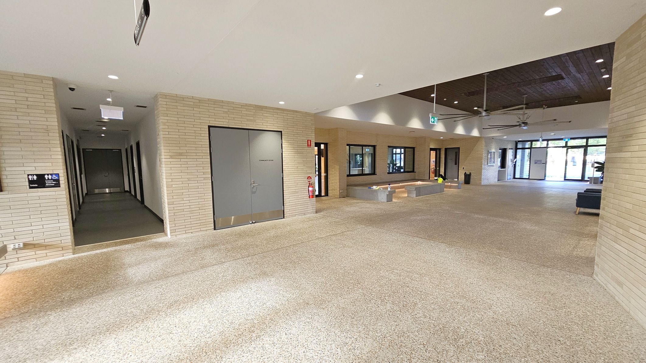 A spacious indoor area with light-coloured brick walls and a speckled floor. On the left, a corridor leads to several doors, with a sign indicating restrooms. In the centre, a large grey door labelled "Community Store" is flanked by a fire extinguisher. The right side features a seating area with benches and a few tables. The ceiling has recessed lighting and ceiling fans. Large windows and glass doors at the far end allow natural light to enter.