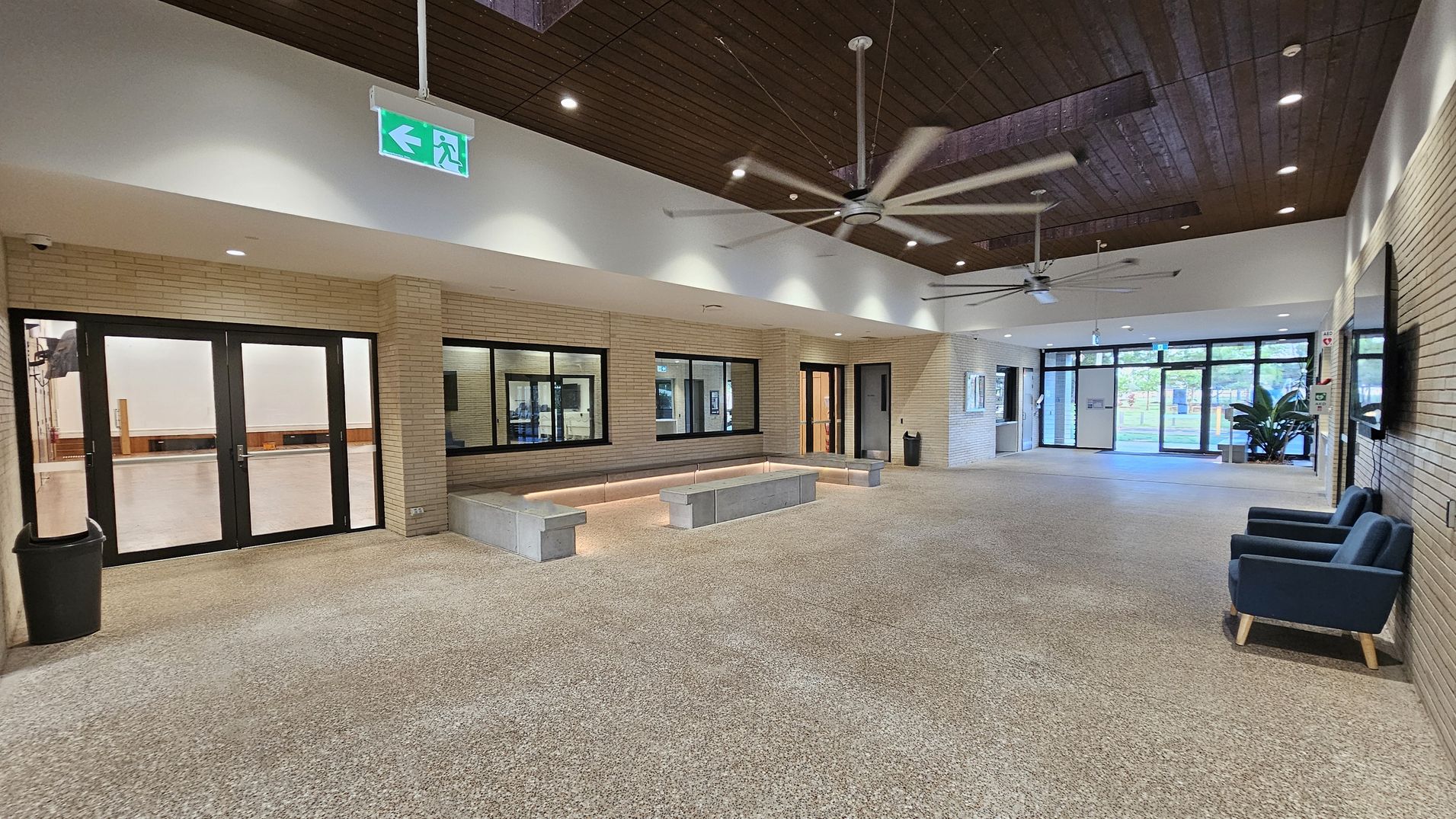 Spacious interior with a terrazzo floor and a wooden ceiling featuring large ceiling fans. On the left, there are black-framed glass doors and windows set in a light brick wall, with a black bin nearby. In the centre, a built-in seating area with concrete benches and soft lighting. On the right, two blue armchairs are placed against the wall, and large glass doors lead outside. An illuminated green exit sign hangs from the ceiling.