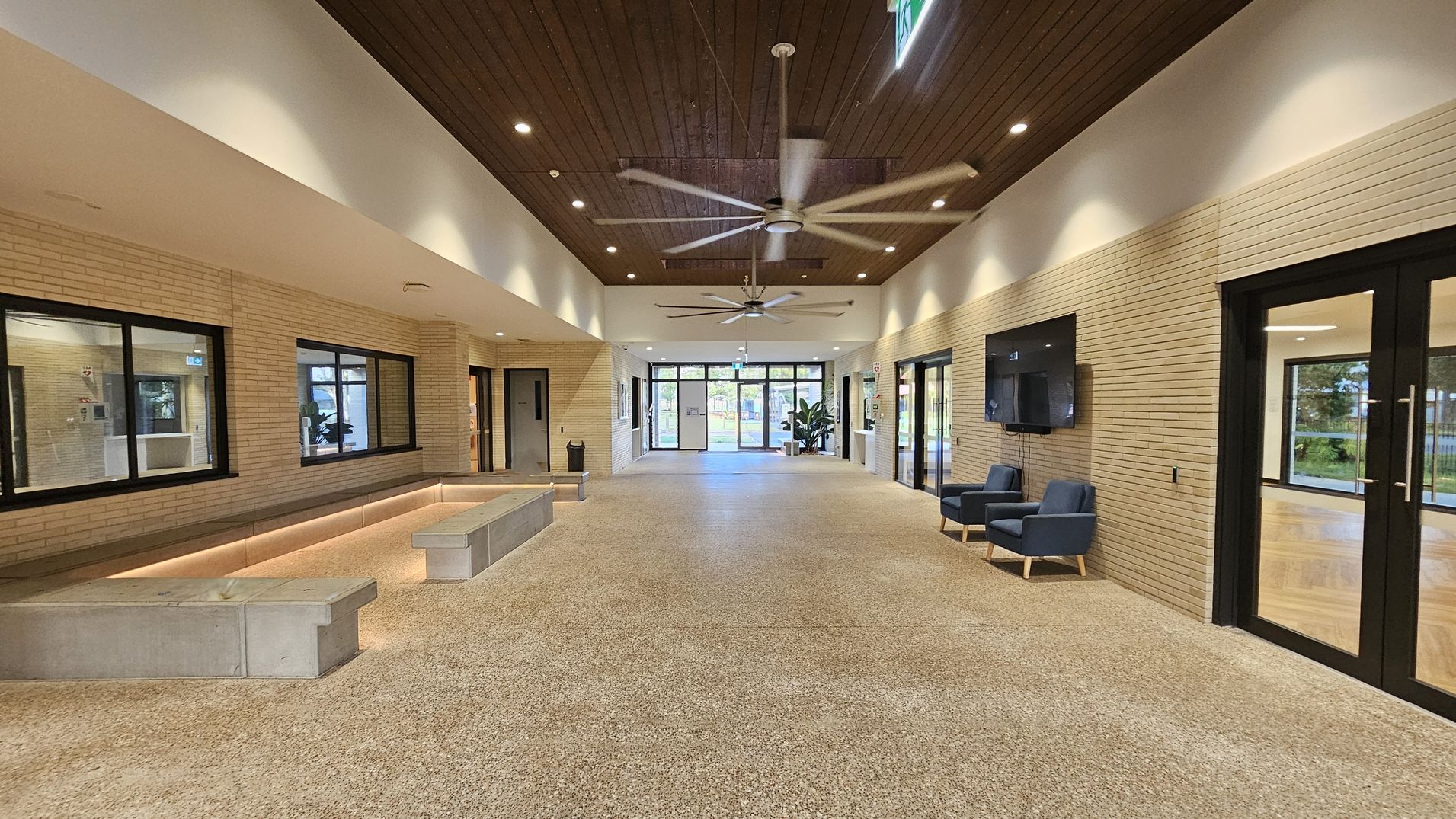 Spacious interior with a polished stone floor and light brick walls. On the left, a long bench with underlighting runs beneath large windows. On the right, two blue armchairs sit below a wall-mounted TV, next to glass doors. The ceiling is wooden with recessed lighting and two large ceiling fans. At the far end, glass doors lead outside, flanked by potted plants.