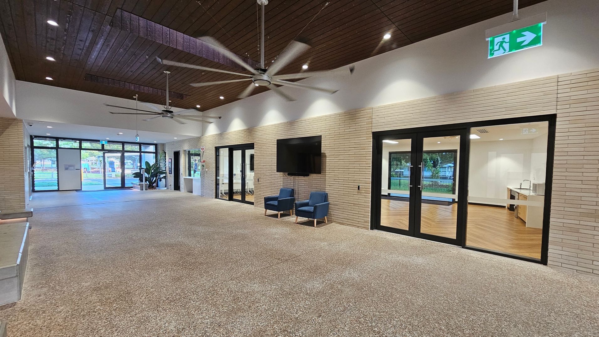 A spacious interior with a polished stone floor and a wooden ceiling featuring recessed lighting and large ceiling fans. On the right, there are glass doors leading to a room with wooden flooring and a kitchenette. Two blue chairs sit beneath a wall-mounted TV. On the left, large windows and doors provide a view of greenery outside. An emergency exit sign is visible above the doors.