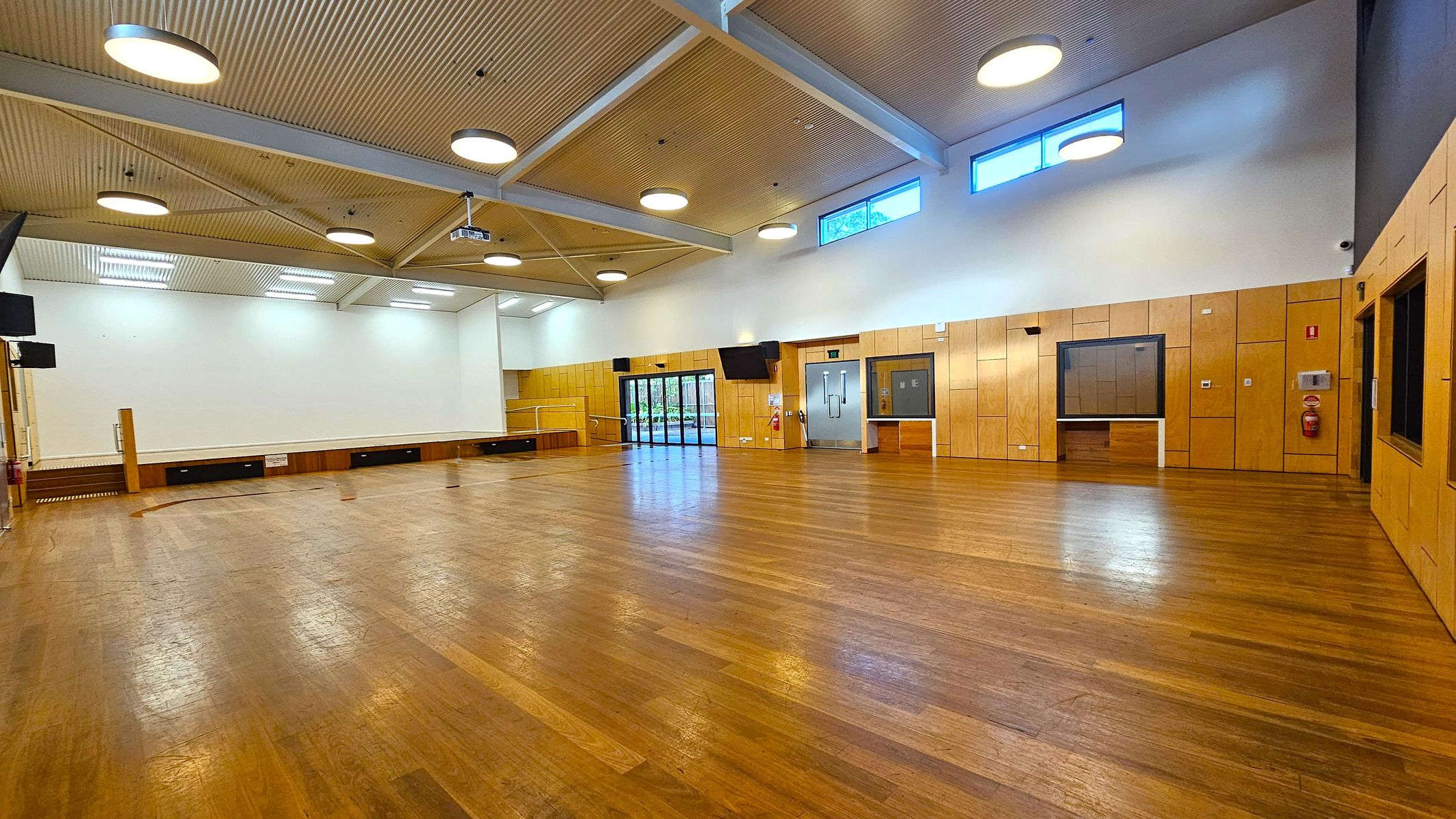 A spacious hall with a polished wooden floor and a high ceiling featuring circular lights. The left side has a stage area with a white backdrop and wooden panelling. On the right, there are large windows and a set of double doors. The walls are lined with light wood panels, and there are several wall-mounted screens. The ceiling has exposed beams and additional lighting fixtures.
