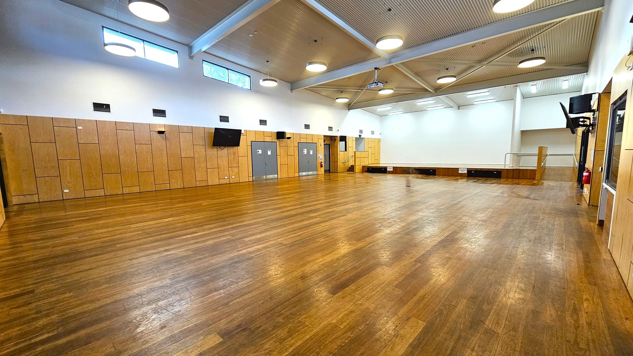 A spacious hall with a polished wooden floor and a high ceiling featuring exposed beams and circular lights. The walls are lined with light wood panelling and have several grey doors. Two large screens are mounted on the walls. In the background, there is a small stage area with steps leading up to it. High windows near the ceiling allow natural light to enter. A fire extinguisher is visible on the right side.