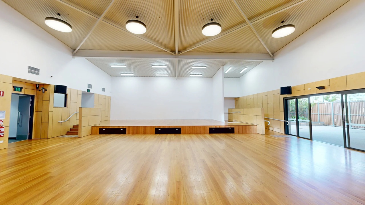 A spacious room with a wooden floor and a high, light-coloured ceiling featuring circular lights. In the centre is a raised stage with a wooden front and black panels. On the left, there is a door and a small staircase leading to the stage. On the right, large glass sliding doors open to an outdoor area with a wooden fence. The walls are white with light wood panelling on the lower half.
